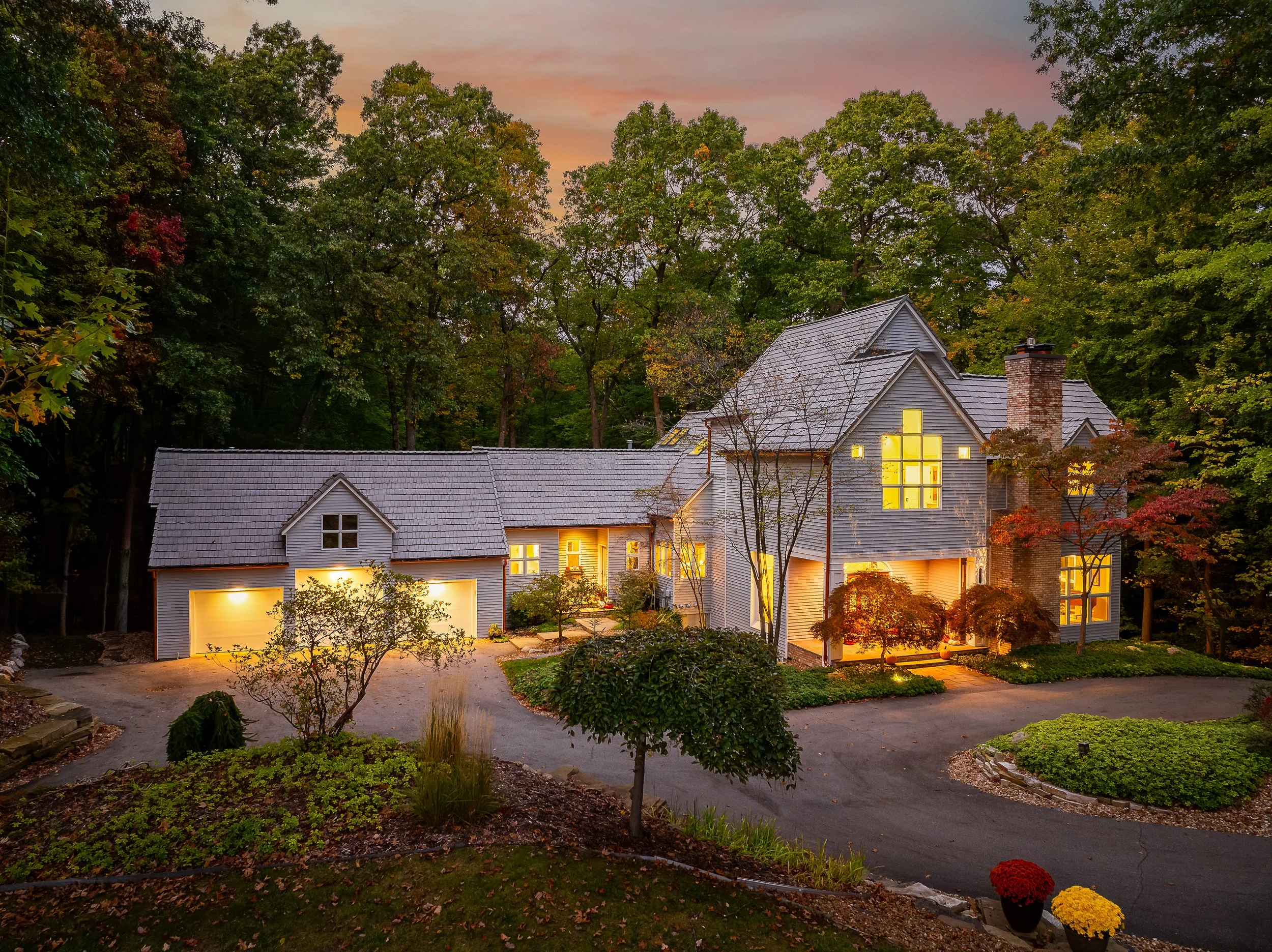 A multi-story house illuminated at dusk, surrounded by trees with fall foliage, featuring large windows, a brick chimney, and a curved driveway with landscaping.