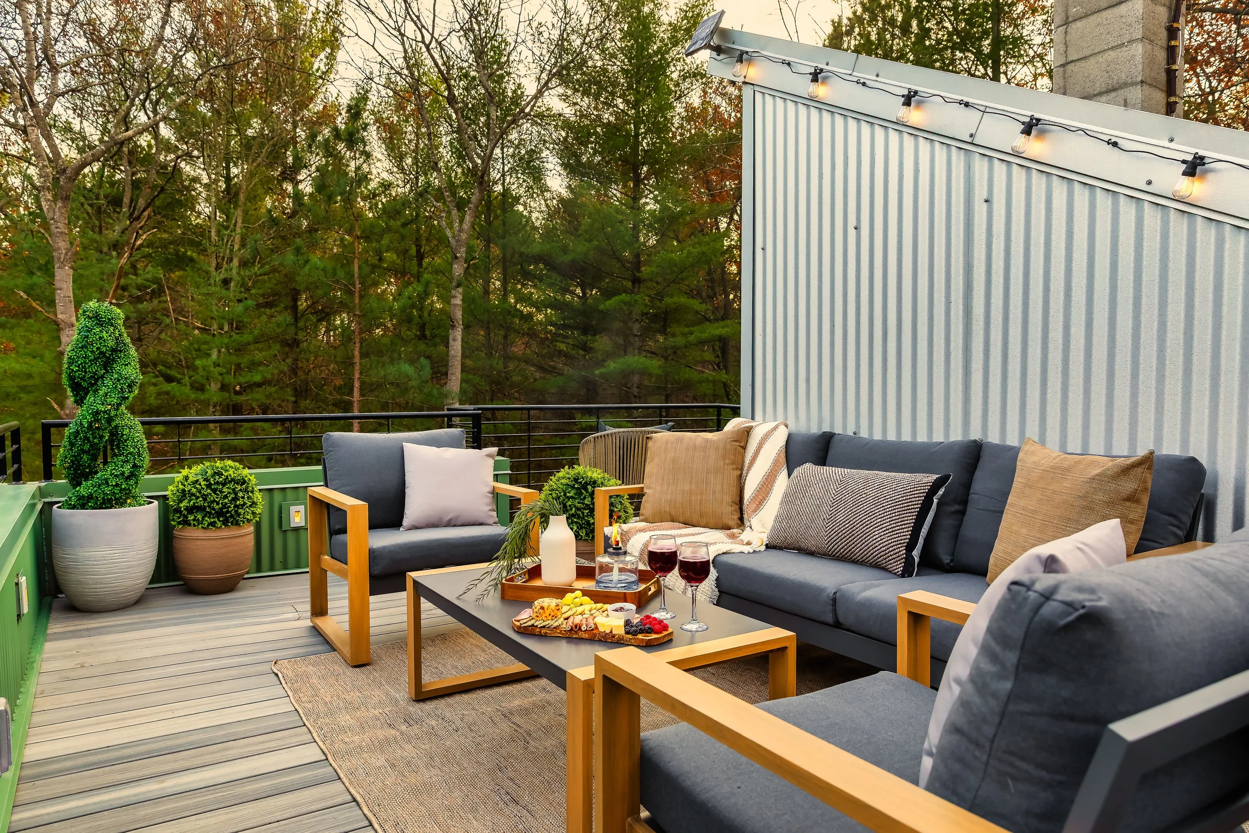 Outdoor rooftop patio with gray and beige seating, decorated with pillows, a coffee table with wine and snacks, and potted plants, surrounded by trees and string lights.