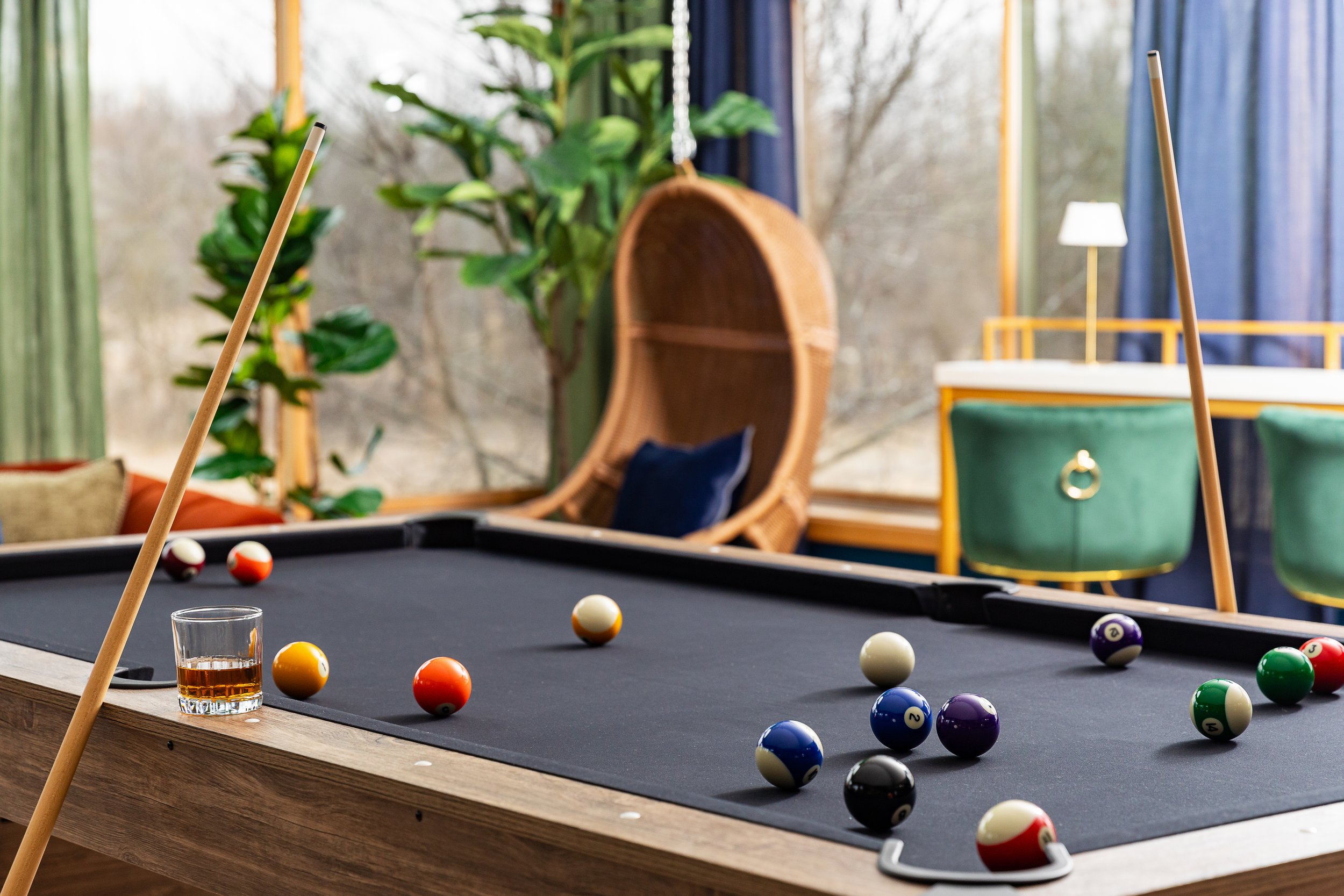 A pool table with several pool balls scattered on the surface, a glass of dark beverage on one corner, in a room with lush green plants, a woven hanging chair with a cushion, and large windows with blue curtains.