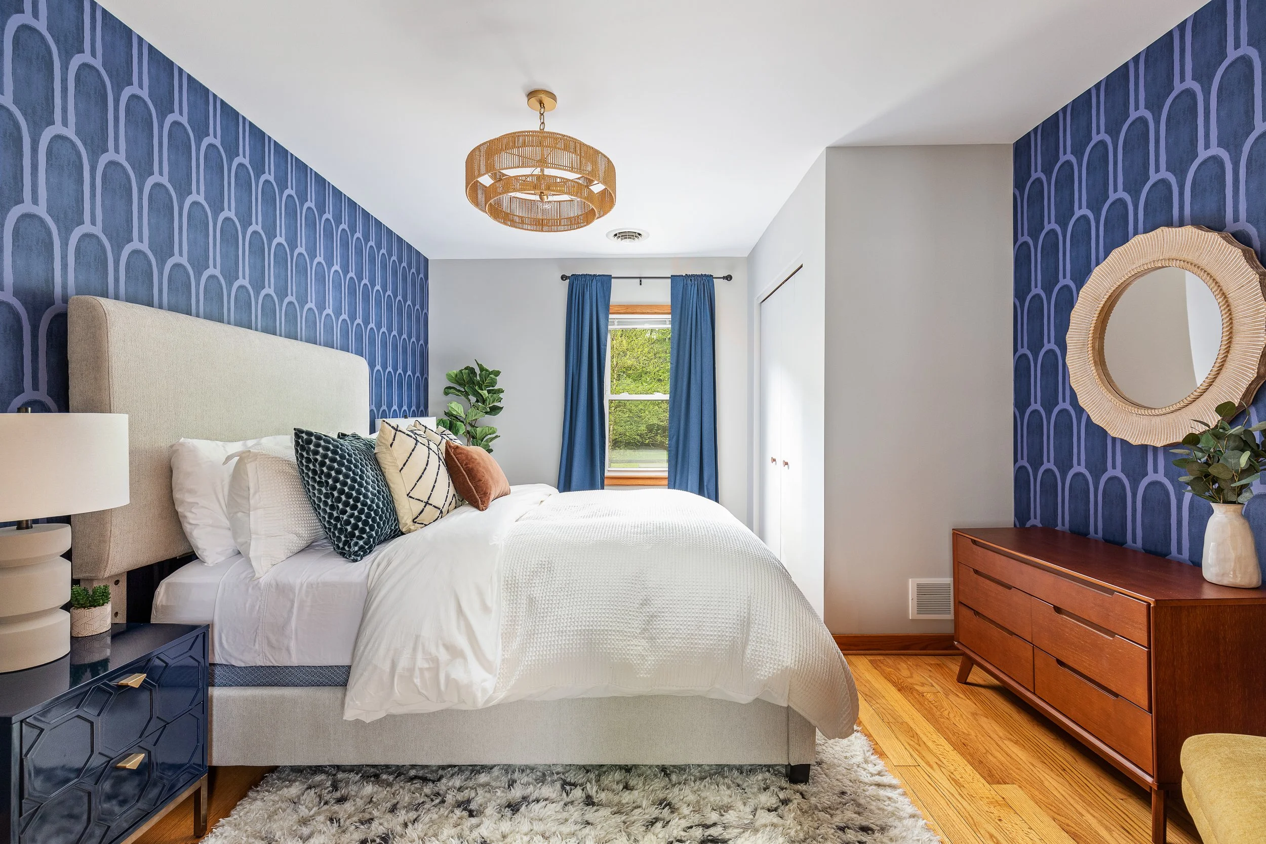 A bedroom with a white bed, blue accent wall with pattern, wooden dresser, and a window with blue curtains.
