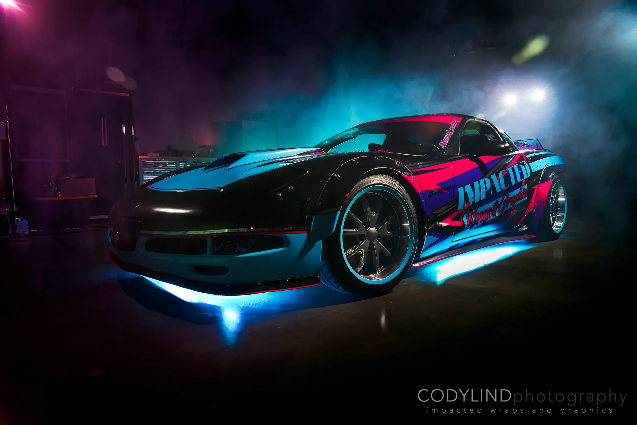 A black sports car with vibrant blue and pink racing graphics, illuminated with glow lights underneath, in a dark workshop or photo studio with colorful lighting effects.