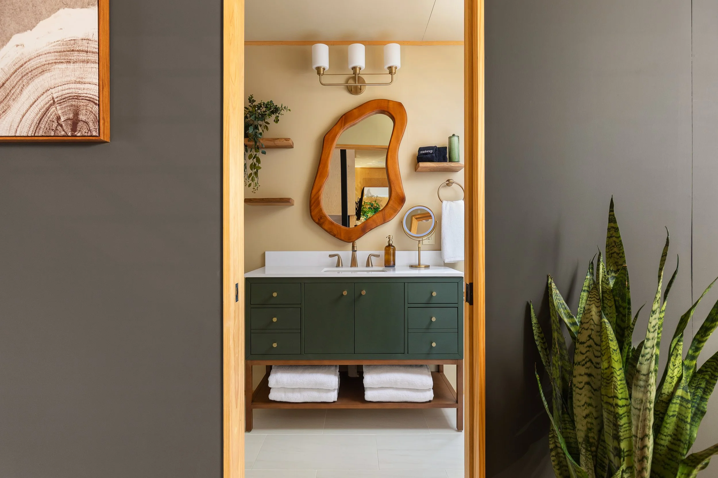 View of a bathroom vanity with a green cabinet, white countertop, oval mirror with a wooden frame, wall-mounted light fixture, and a small round mirror, with shelves holding toiletries, and a towel hanging on a ring, with a plant on the right side.