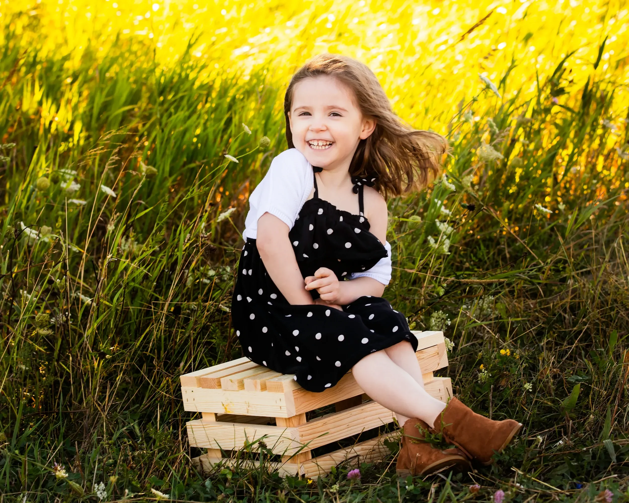 A smiling young girl with brown hair, wearing a black polka dot dress over a white shirt and brown boots, sitting on a wooden crate in a field of tall grass and wildflowers during sunset.