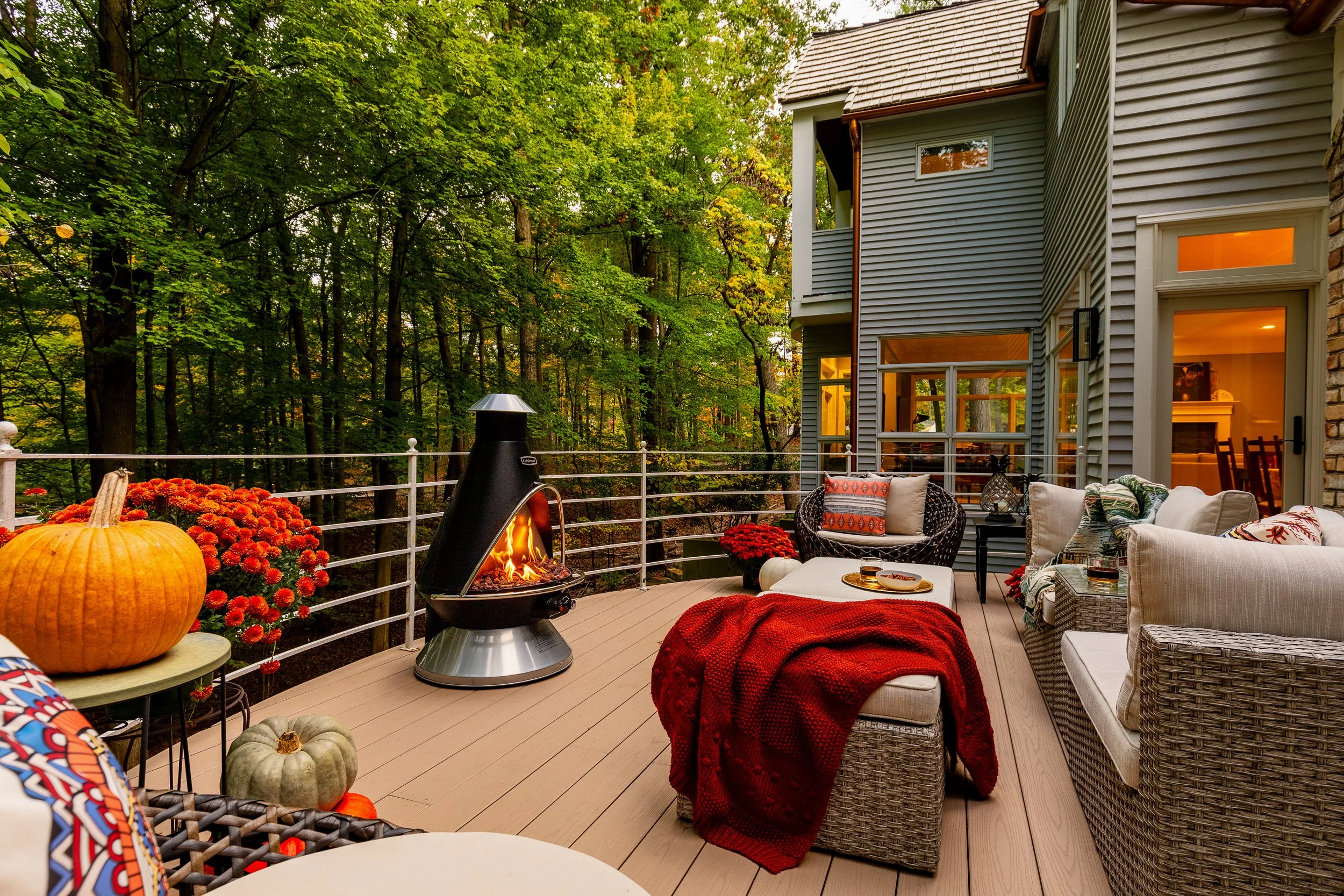 A cozy outdoor patio deck with wicker furniture, a red blanket, pumpkins, a fireplace, and fall decorations, surrounded by green trees.