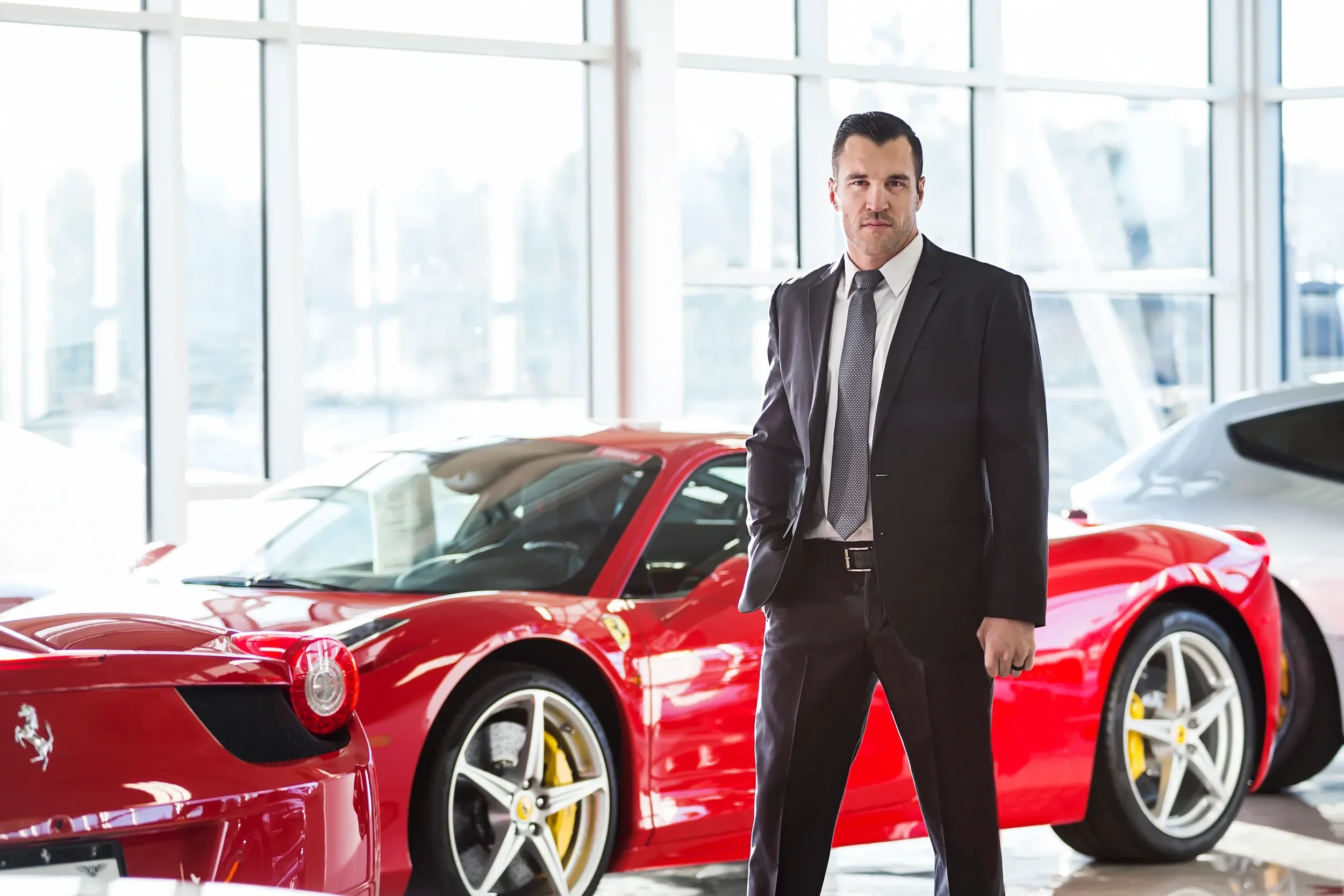 A man in a black suit stands with his hand in his pocket in front of a red sports car inside a car dealership with large windows.