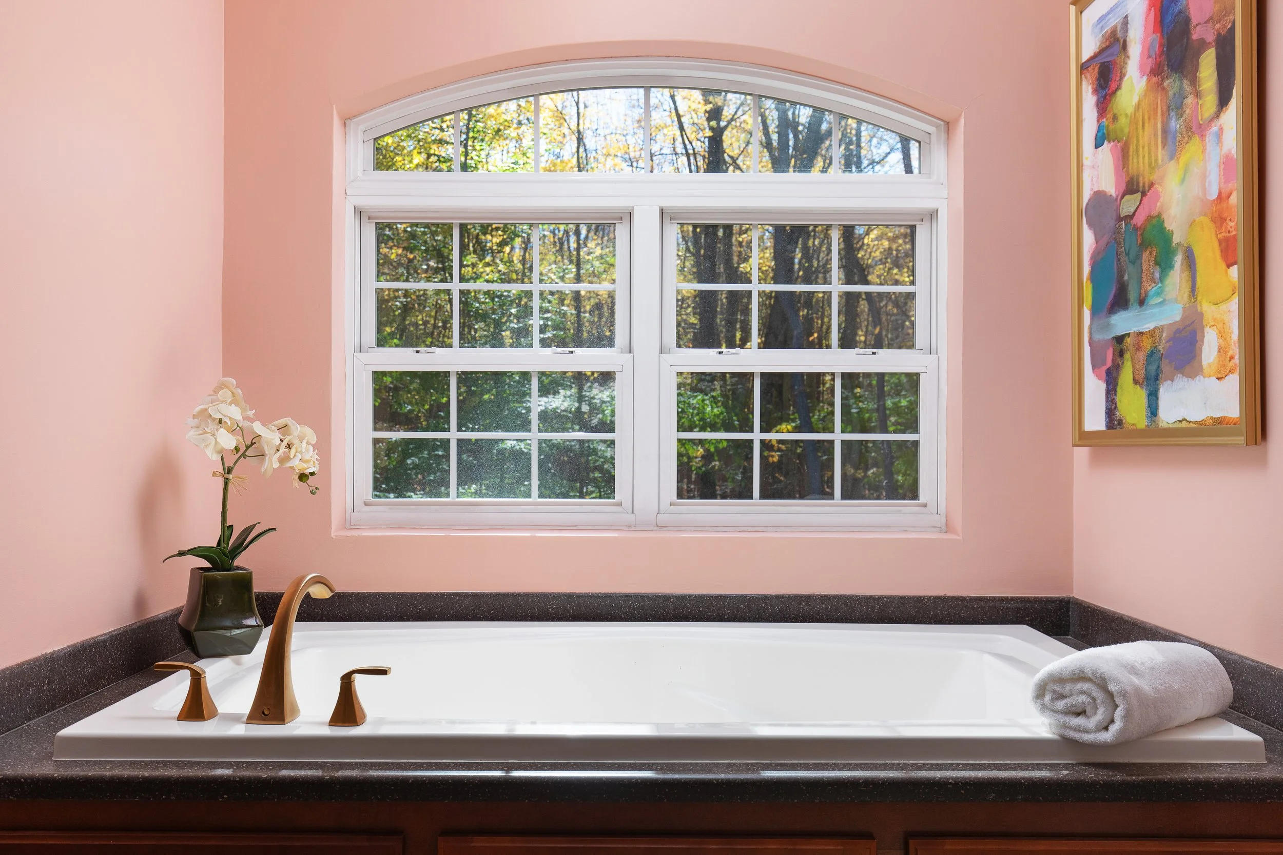 Bathroom with a white bathtub, pink walls, a large window showing trees outside, a white rolled towel on the edge of the tub, a vase with white orchids, and colorful abstract artwork on the wall.