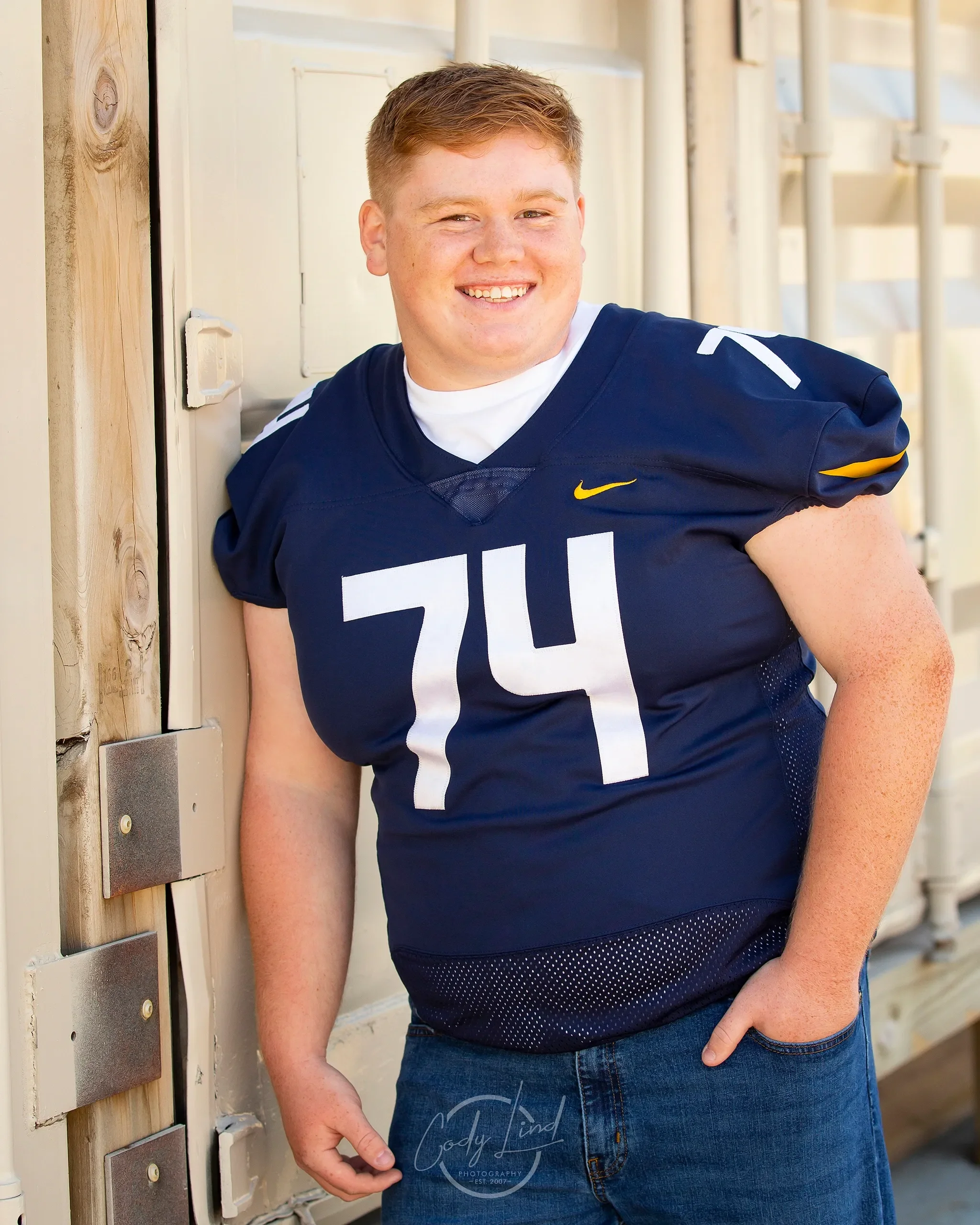 A young man in a navy football jersey with the number 74, smiling and leaning against a wooden wall.