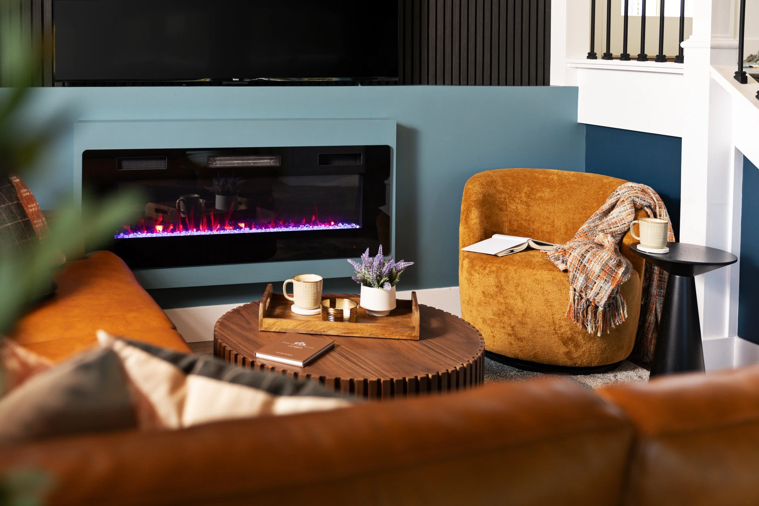 Living room with a modern electric fireplace, a round wooden coffee table with a small book, a tray with a mug and a small plant, a mustard-colored armchair with a blanket and a mug, a side table with a mug, and a brown sofa in the foreground.