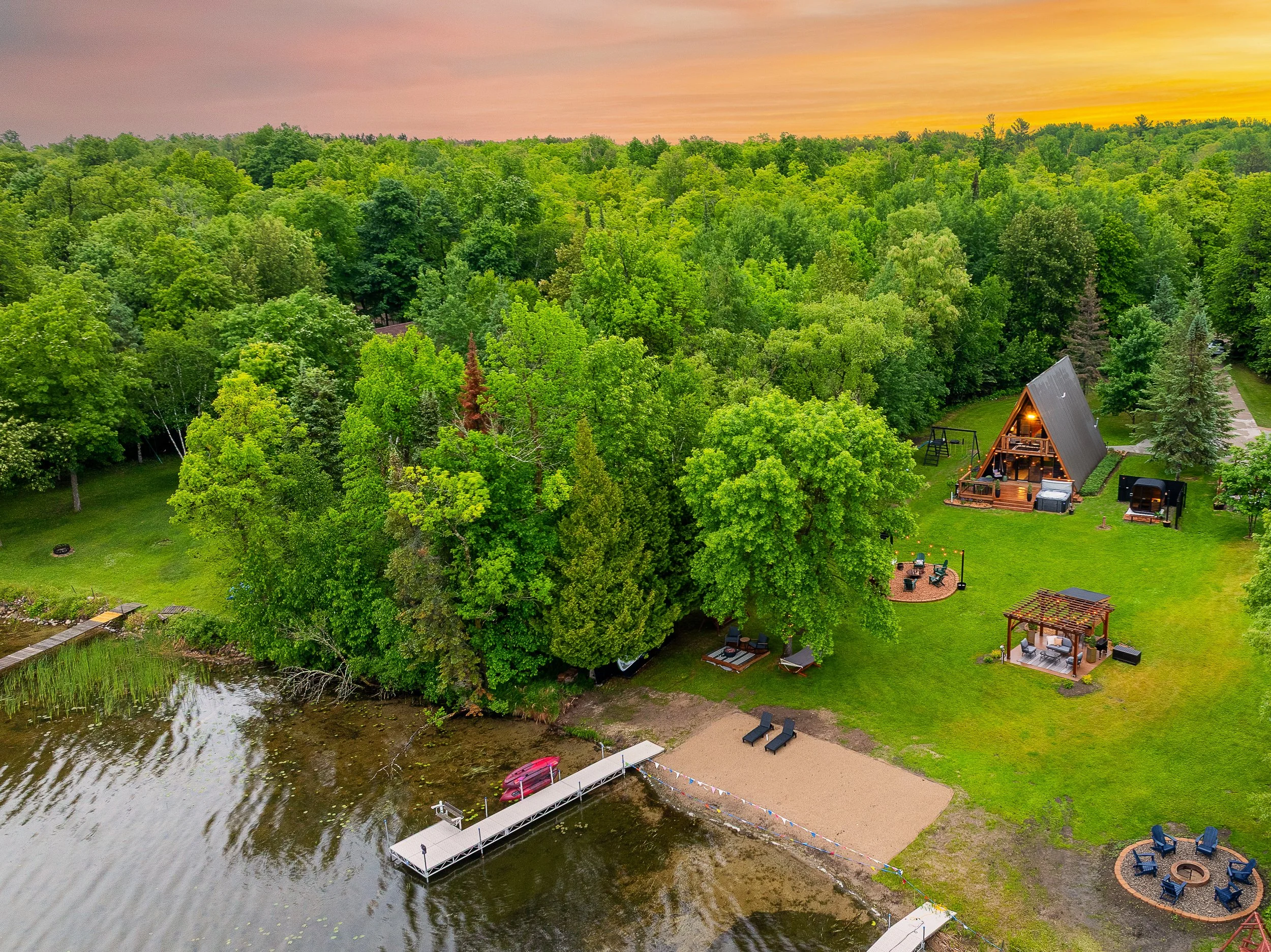 Aerial view of a lakeside property with lush green trees, a sandy beach area with lounge chairs and a dock with kayaks, a large A-frame house with outdoor seating, a smaller gazebo, and a fire pit with chairs at sunset.