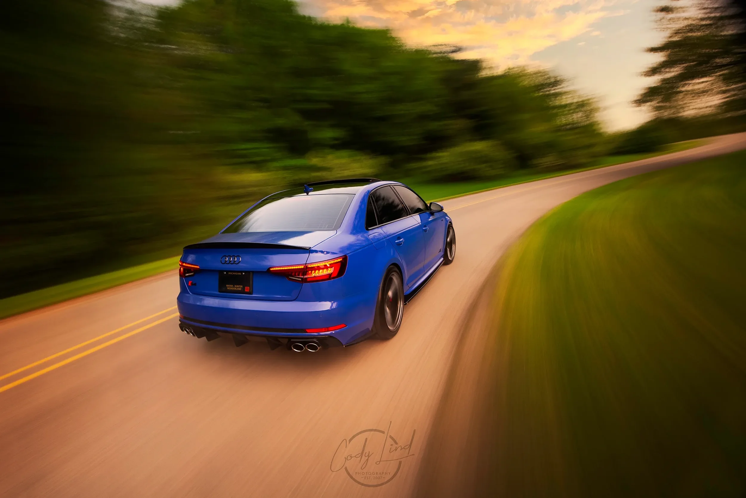 Blue Audi sedan driving on a curved country road during sunset, surrounded by green trees and grass.