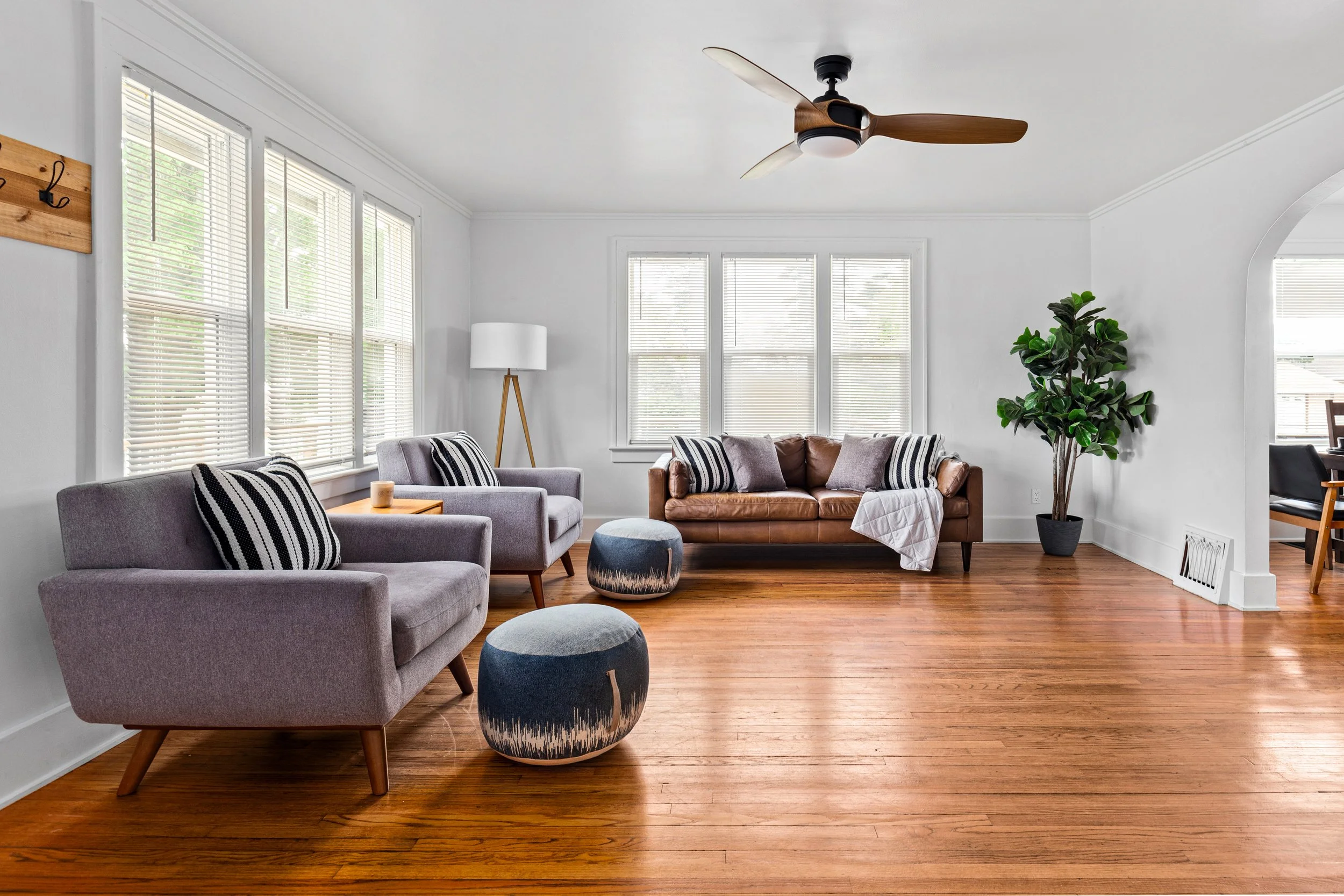 Bright living room with hardwood floors, gray and brown sofas, striped pillows, a standing lamp, windows with blinds, a potted plant, and a ceiling fan.