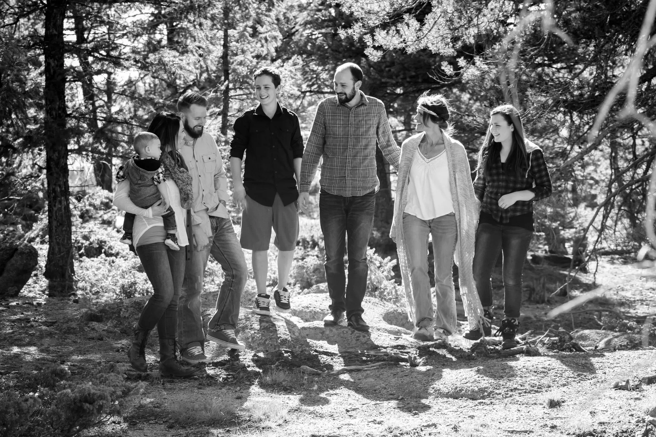 A group of eight people, including a child, walking and smiling in a forest. The group includes men and women of various ages, enjoying an outdoor activity.
