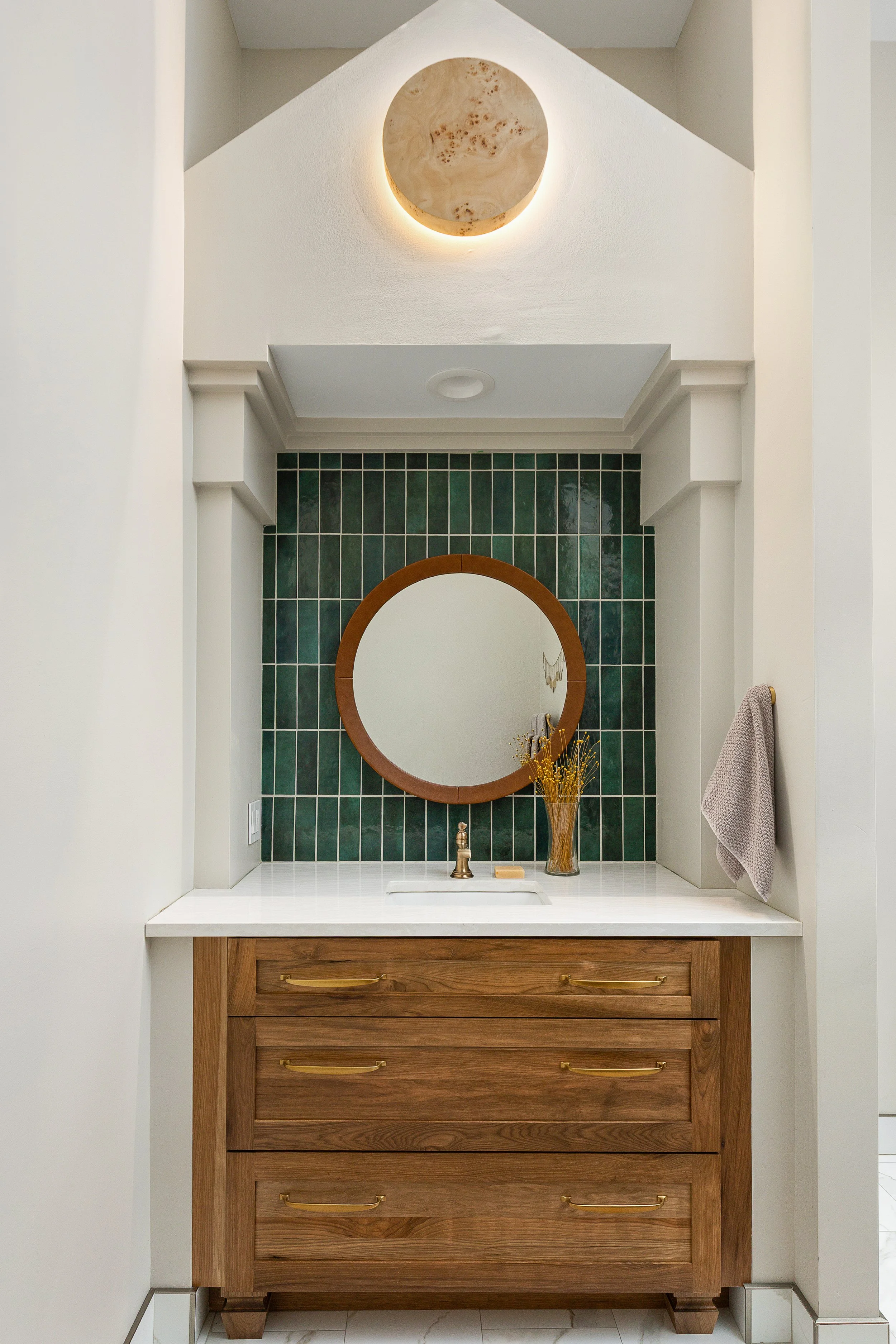 Bathroom vanity with a round mirror, green tiled backsplash, wooden drawers with gold handles, a vase with dried flowers, a soap dish, and a grey towel hanging on the wall.