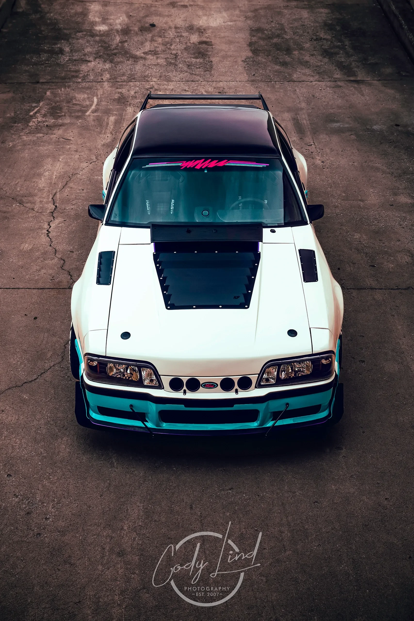 Top view of a modified white and teal race car with black hood vents, front splitters, and a black rear wing, parked on concrete ground. Timefox 2