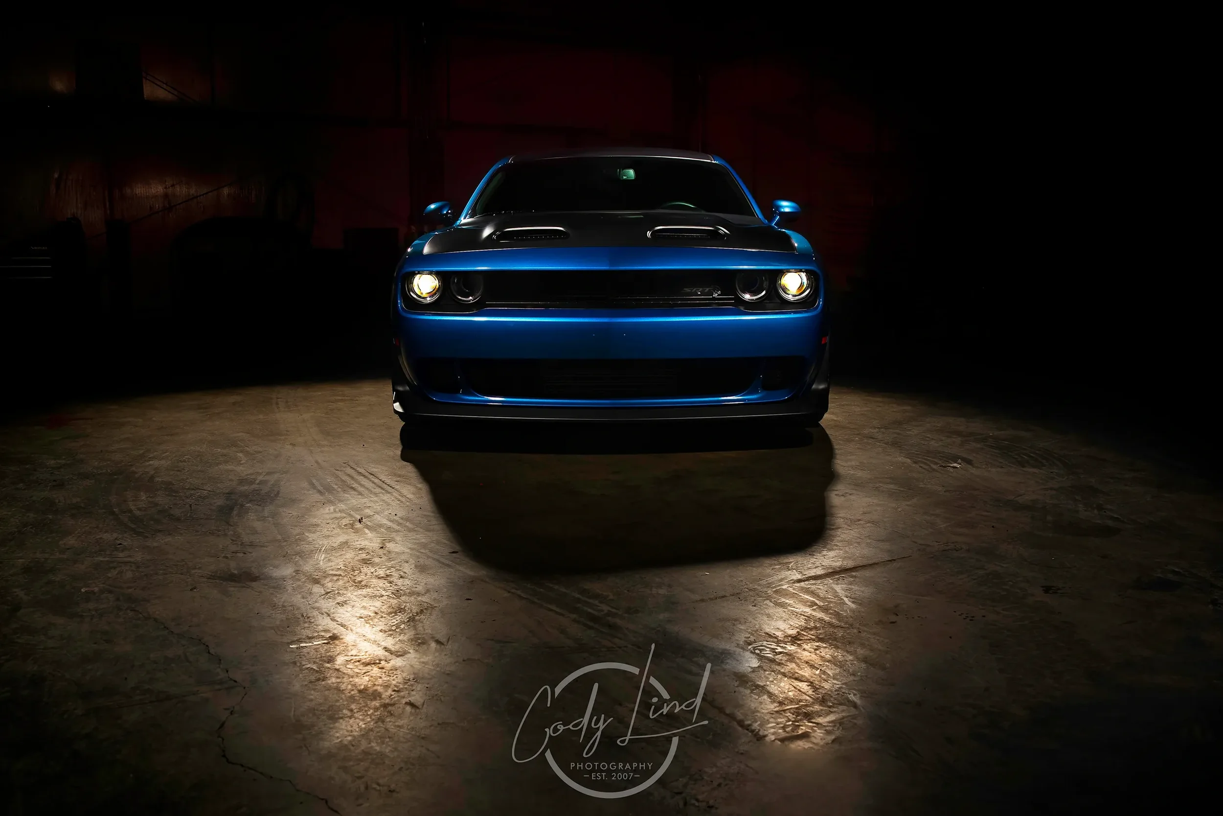 A blue sports car with black hood and hood vents, front view, in a dark warehouse with reflections on the concrete floor.