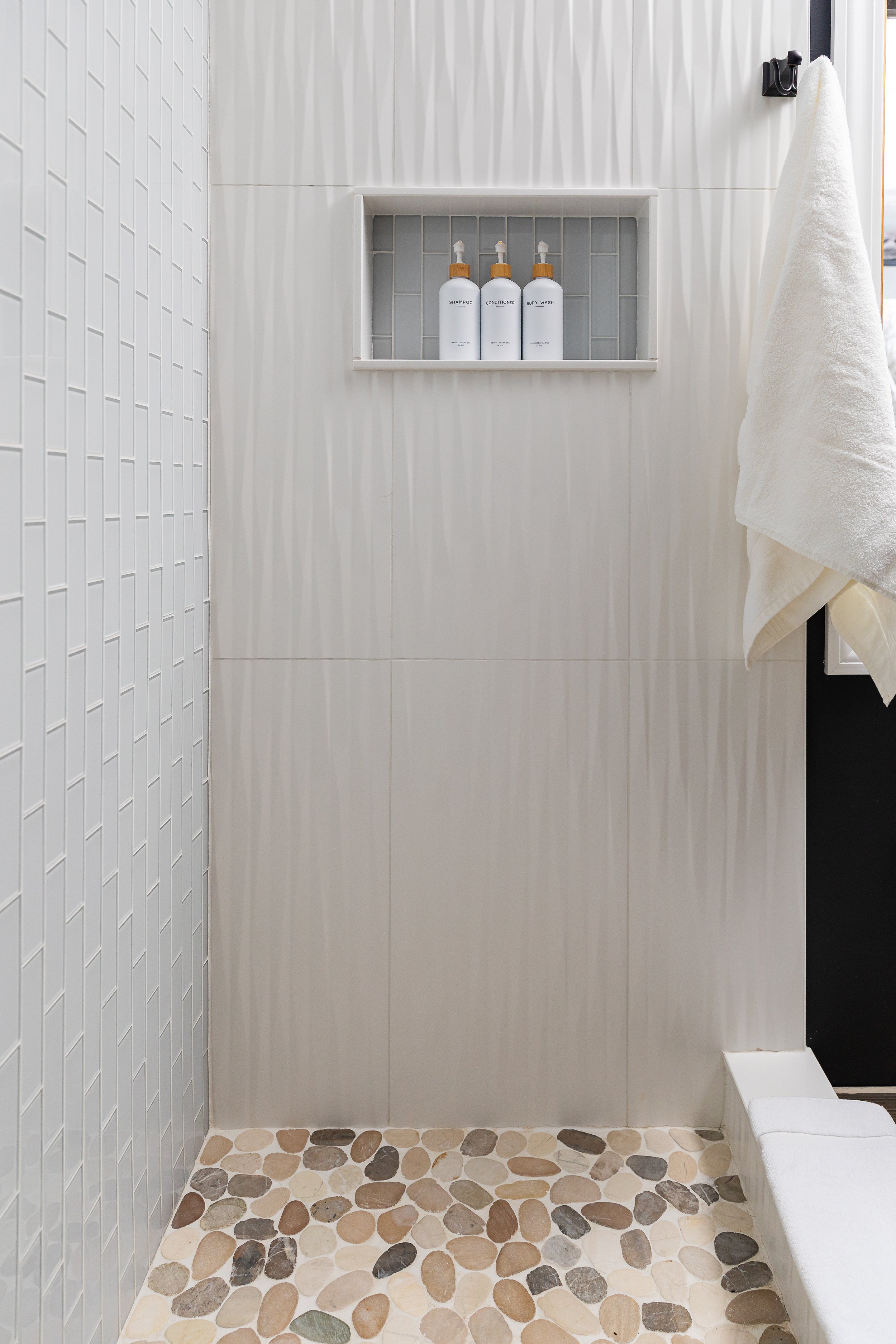 Shower with white tiled wall, shelf with three bottles labeled for shampoo, conditioner, and body wash, white towel hanging on hook, pebble flooring.