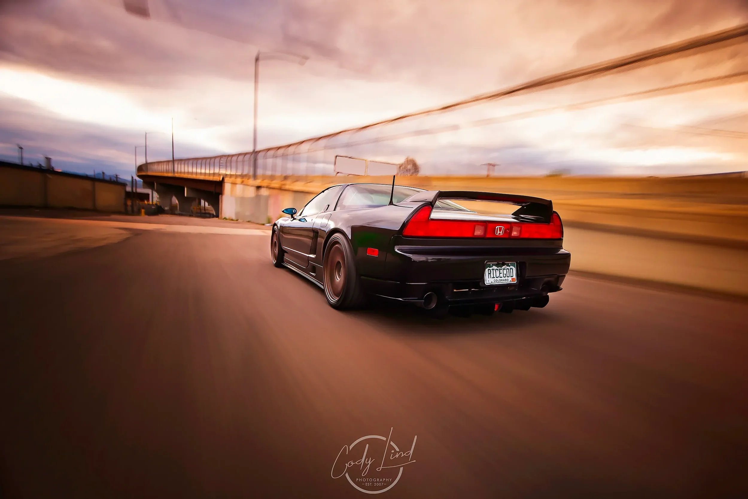 Black Honda sports car with a spoiler driving on a curved road during sunset, with motion blur in the background.