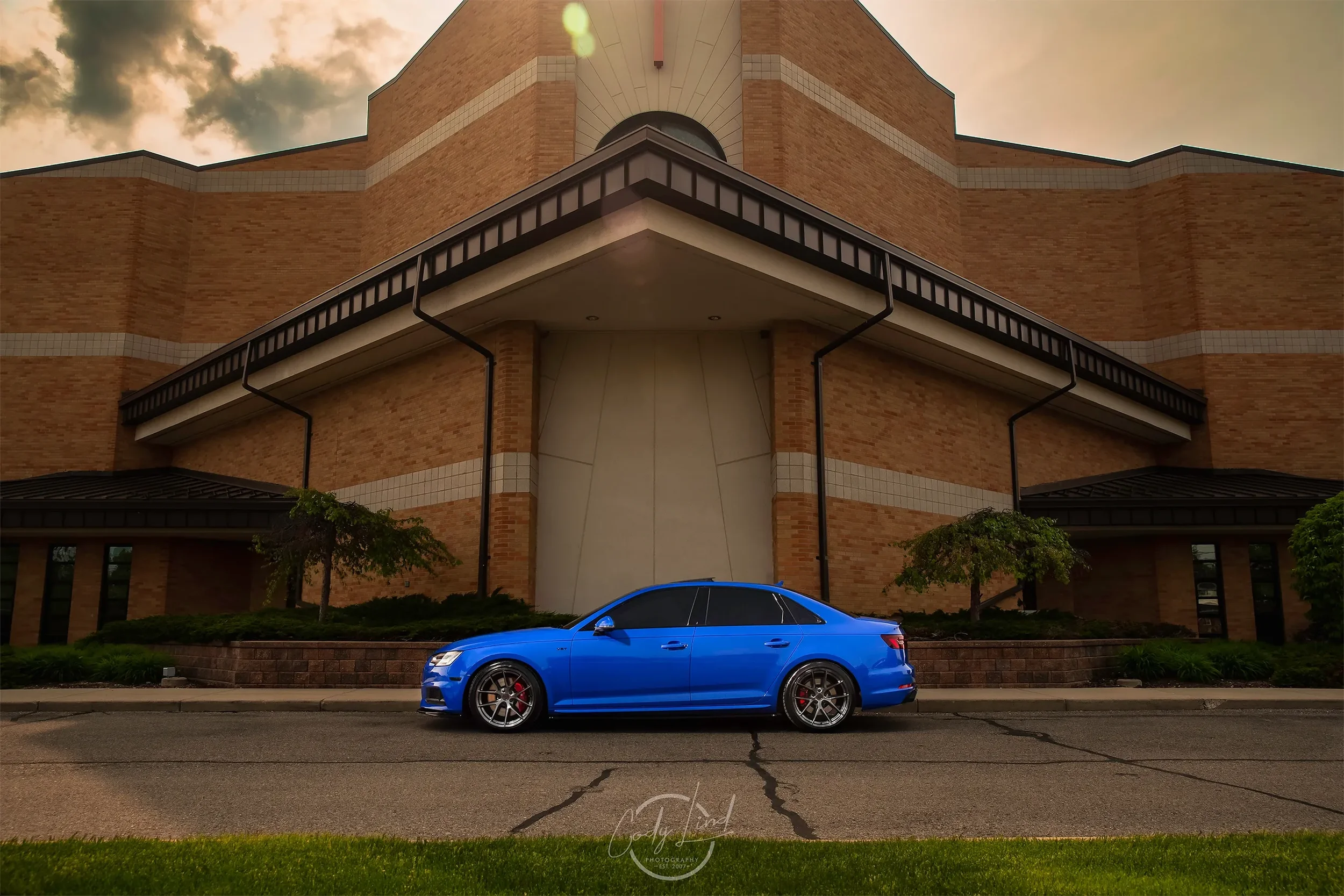 A blue sedan parked on a street in front of a large brick building with trees and landscaped bushes.