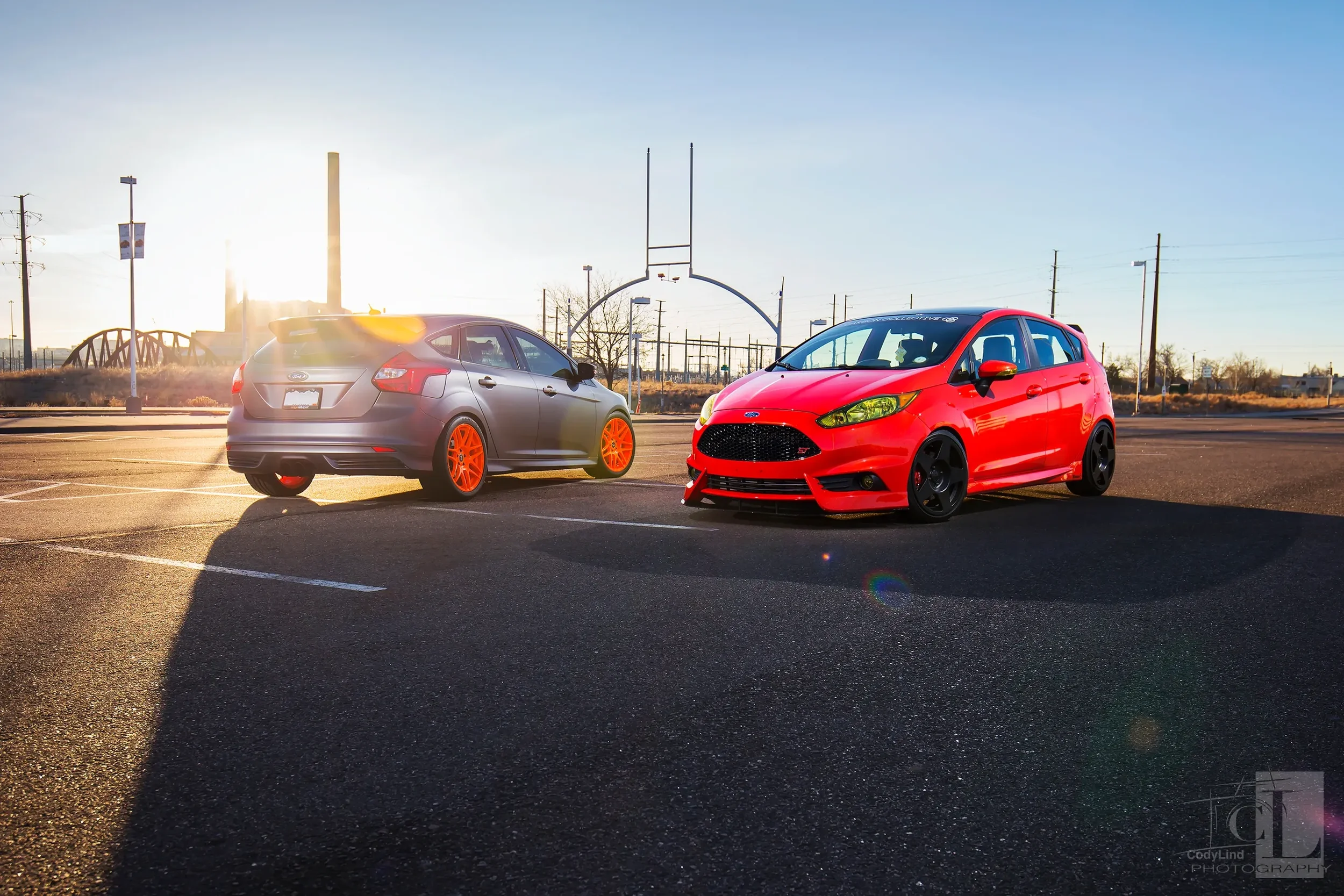 Two modified cars, a gray hatchback with orange wheels and a red hatchback with black wheels, parked in an empty parking lot during sunset with an industrial background.