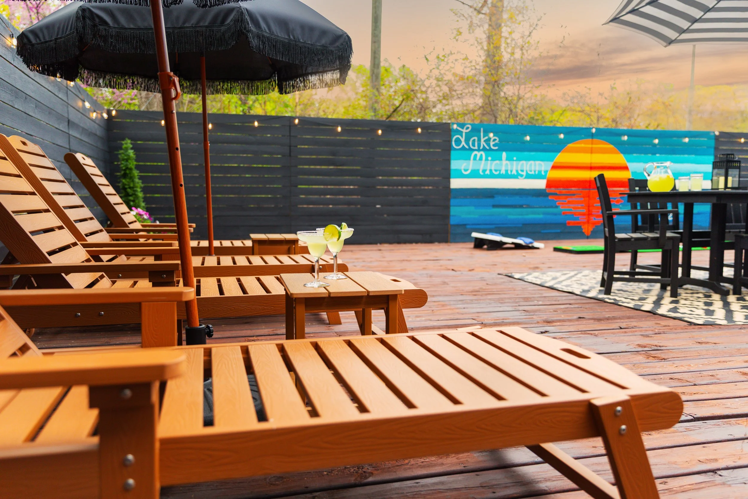 A backyard patio with wooden lounge chairs, umbrellas, and a table with drinks. There is a mural on the fence that says 'Lake Michigan' with a sunset and water design.