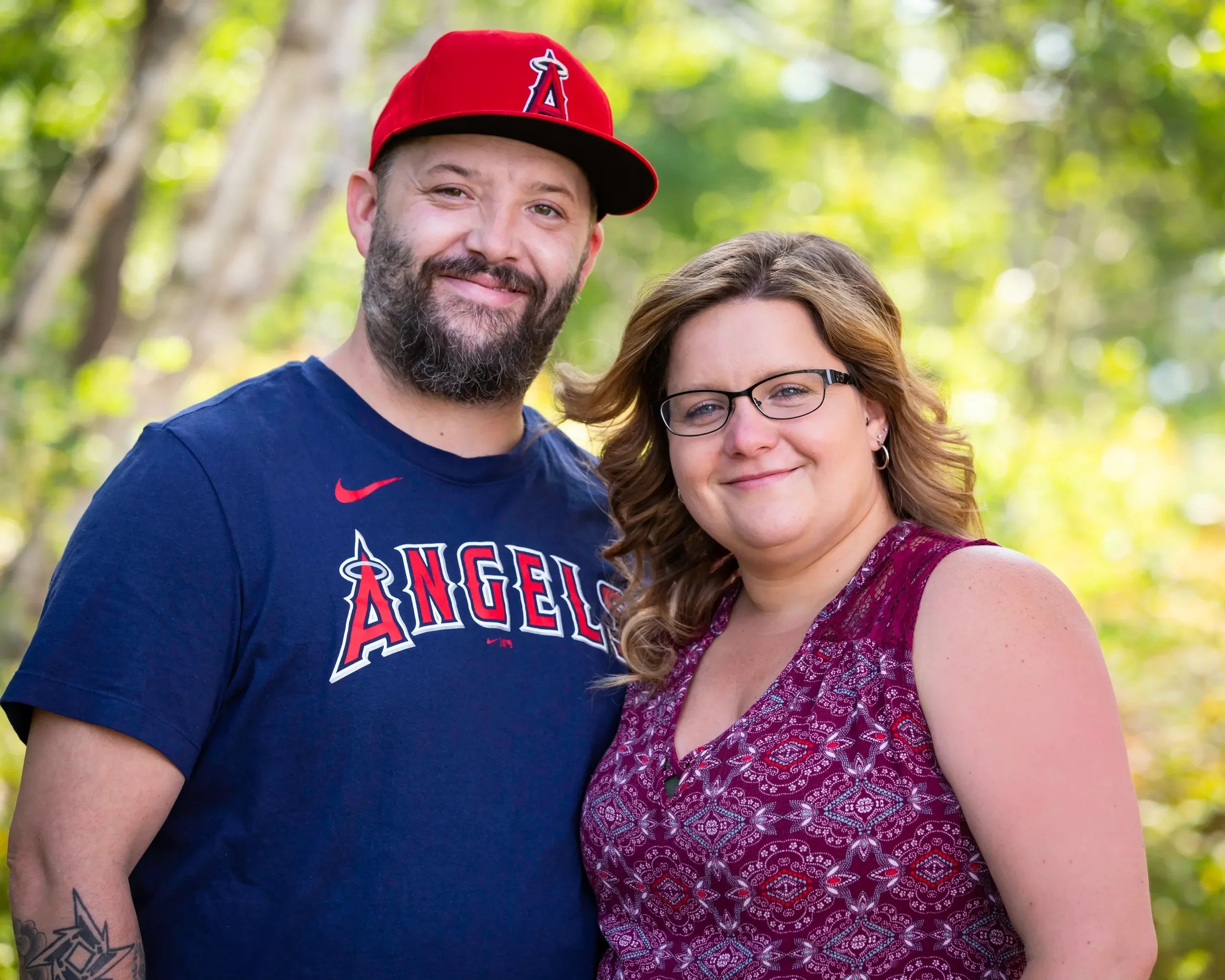 A smiling man with a beard wearing a Los Angeles Angels shirt and red cap, standing next to a smiling woman with glasses and wavy hair, outdoors with green trees in the background.