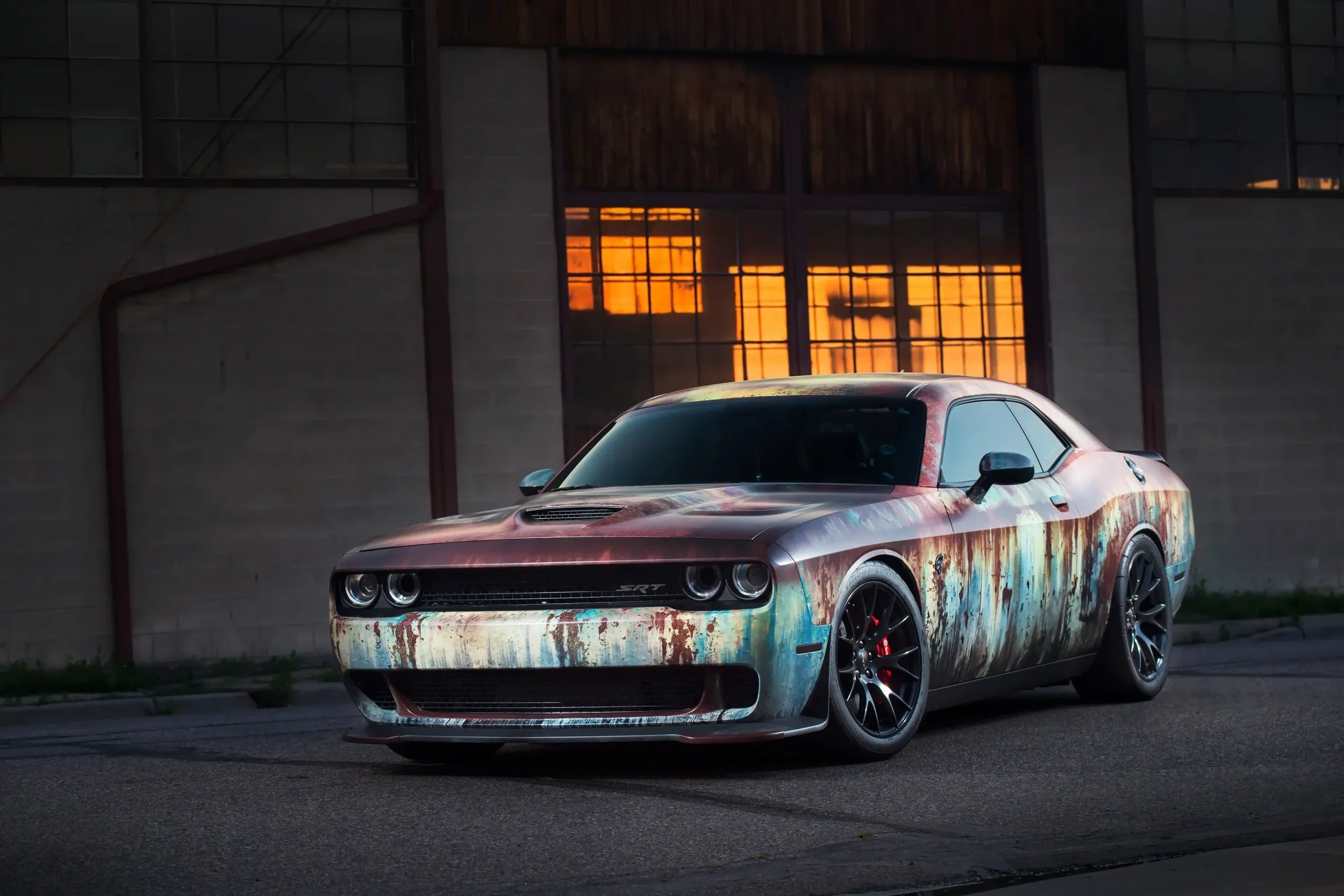 A rusty, weathered Dodge Challenger parked outside in front of a building with large windows reflecting an orange sunset.