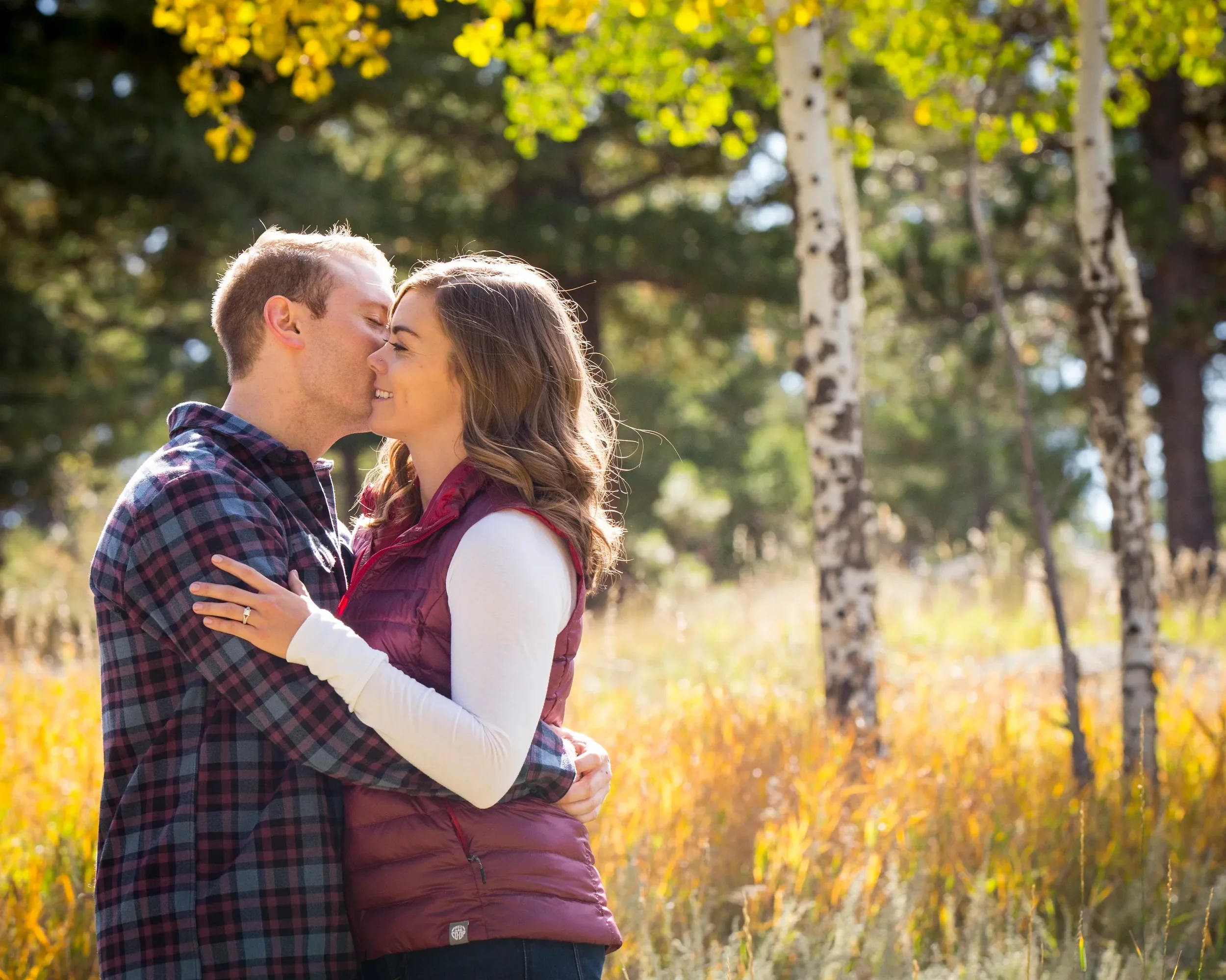 A couple stands close together in a forest with yellow autumn leaves, about to kiss or share an intimate moment.
