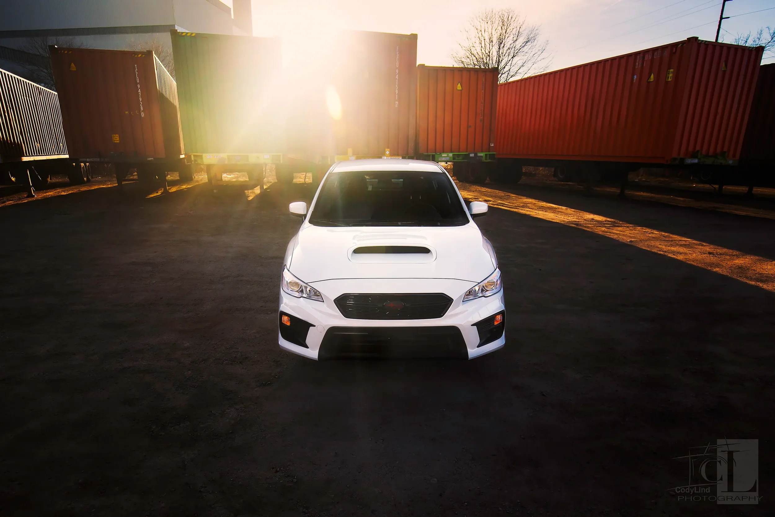 White car parked on black asphalt with cargo containers in the background and sunlight shining behind the containers.