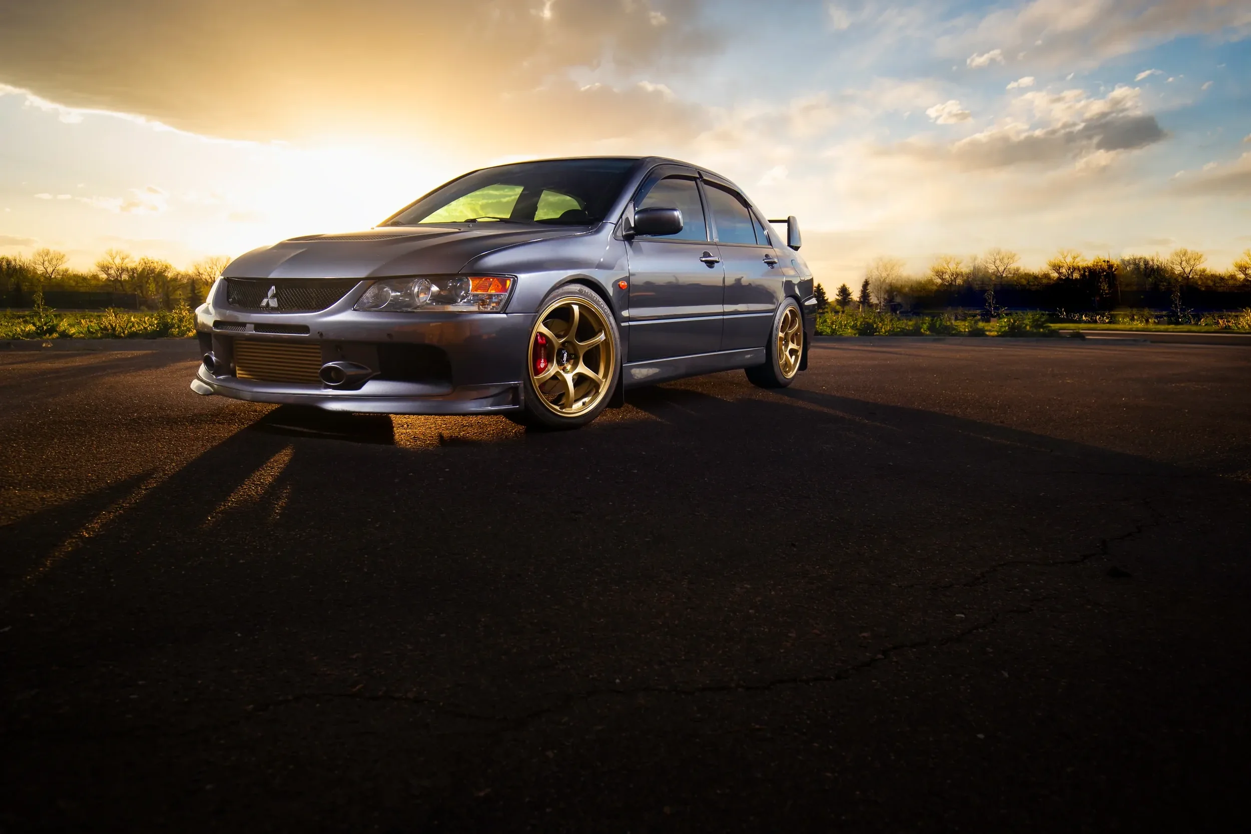 A gray Mitsubishi Lancer Evolution parked outdoors during sunset, with gold racing wheels and a front-mounted intercooler visible.