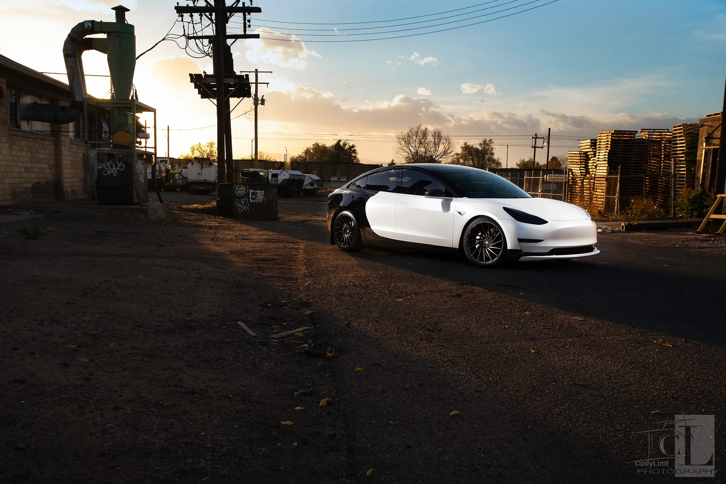 A Tesla Model 3 parked on an industrial lot during sunset, with stacks of pallets and utility poles in the background.