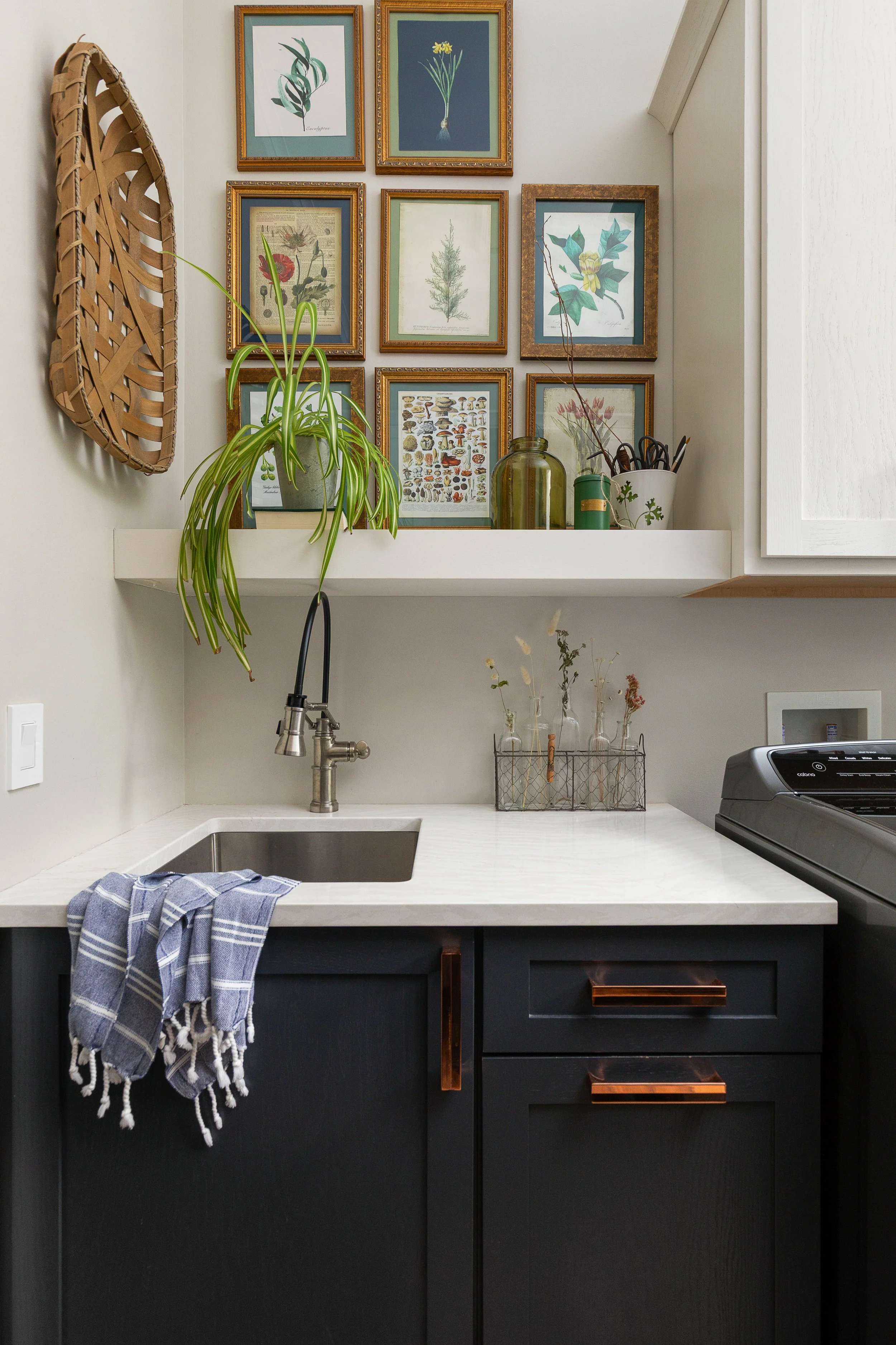 A modern kitchen corner with a black cabinet, white marble countertop, small black sink with a chrome faucet, patterned dish towel hanging from the corner, wall art of framed botanical illustrations above, a potted plant and jars on a white shelf, an