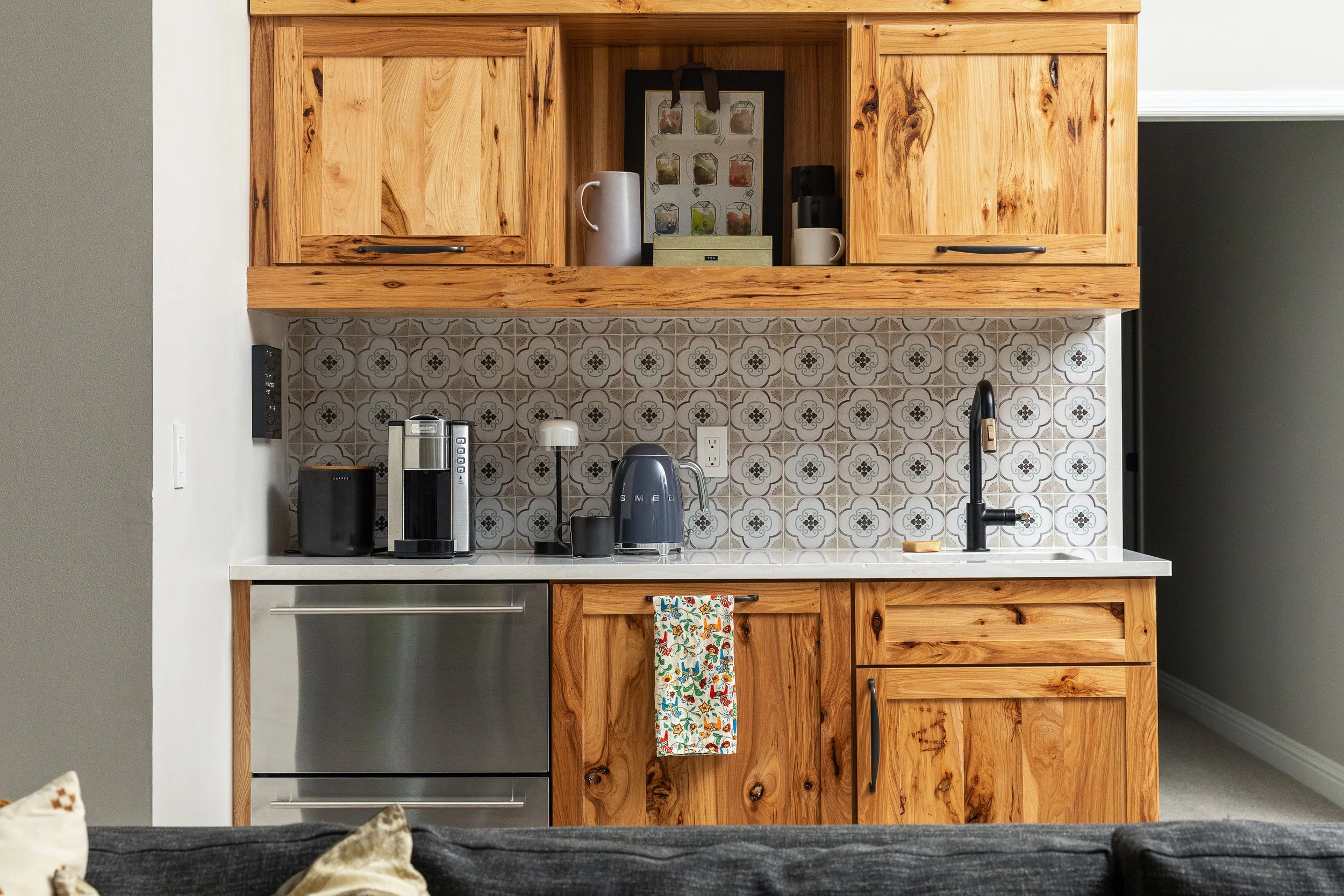 A kitchen with natural wood cabinets, patterned tile backsplash, black faucet, and various appliances including a coffee maker, toaster, and kettle.