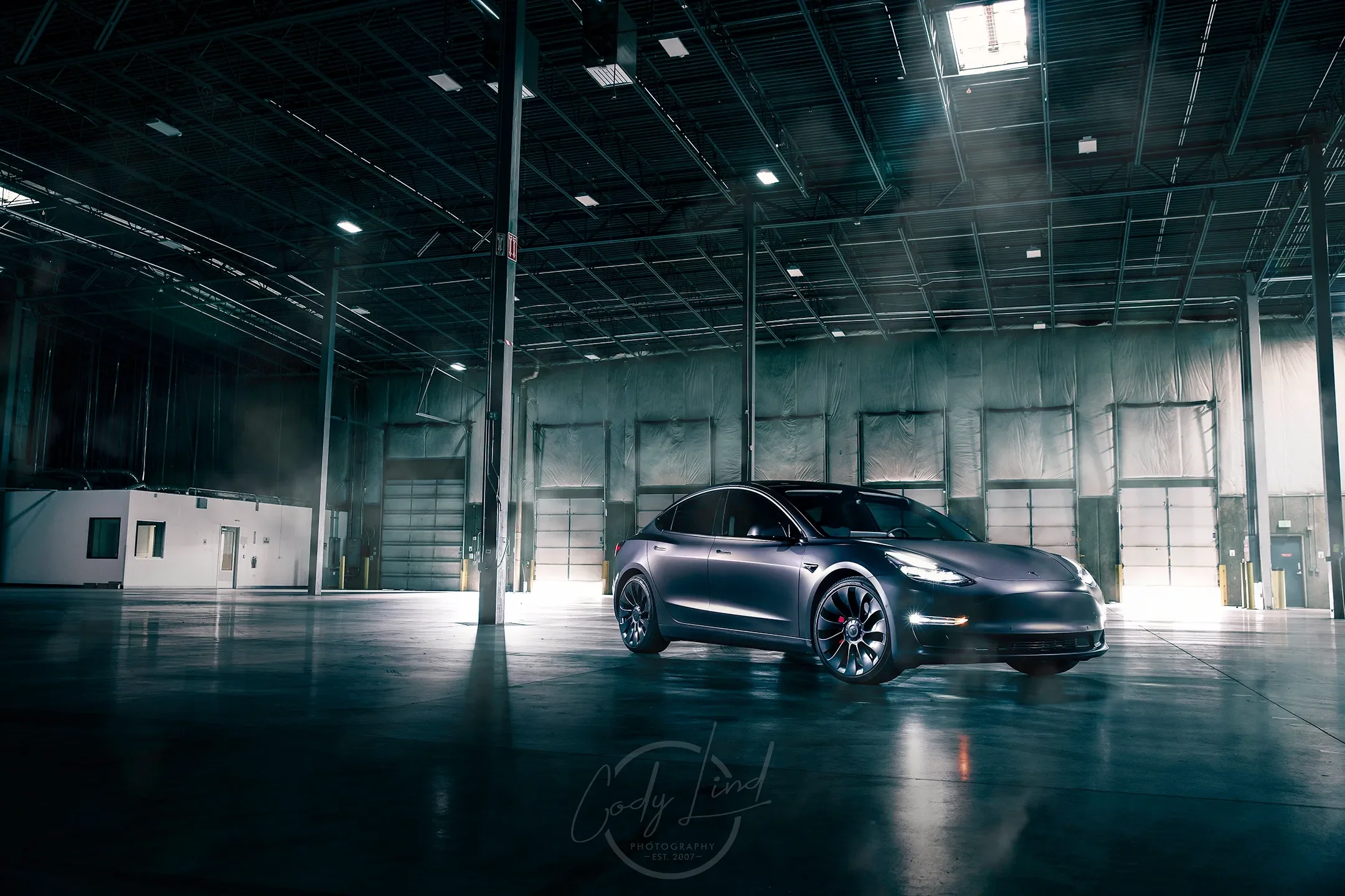 A sleek black Tesla car parked inside a large, empty industrial warehouse with high ceilings and metal beams.