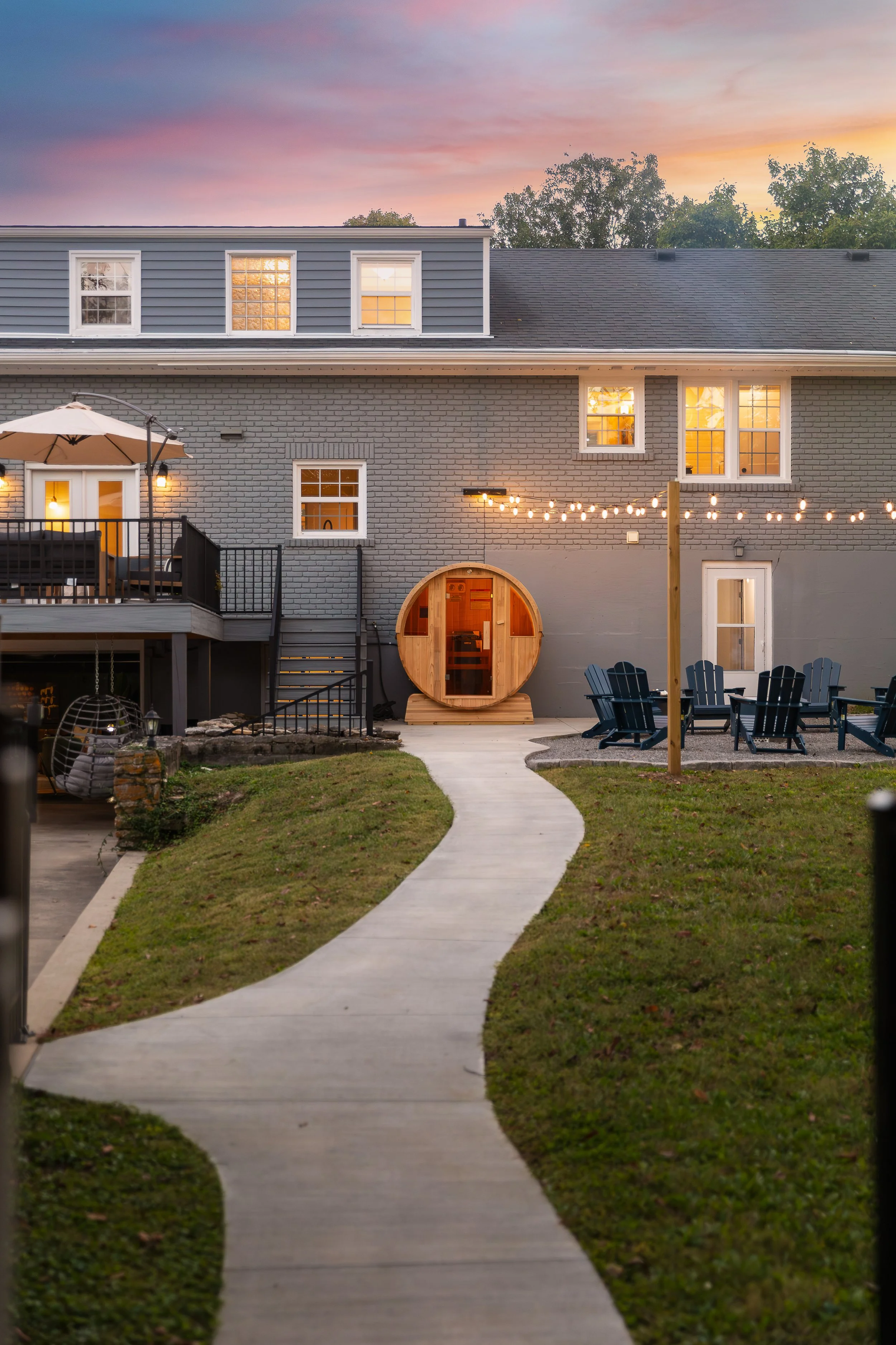 A backyard with a winding concrete pathway leading to a gray brick house at dusk. The house has multiple windows and a small deck with outdoor furniture and an umbrella. A string of lights is hung across the yard, and a wooden barrel sauna is placed 