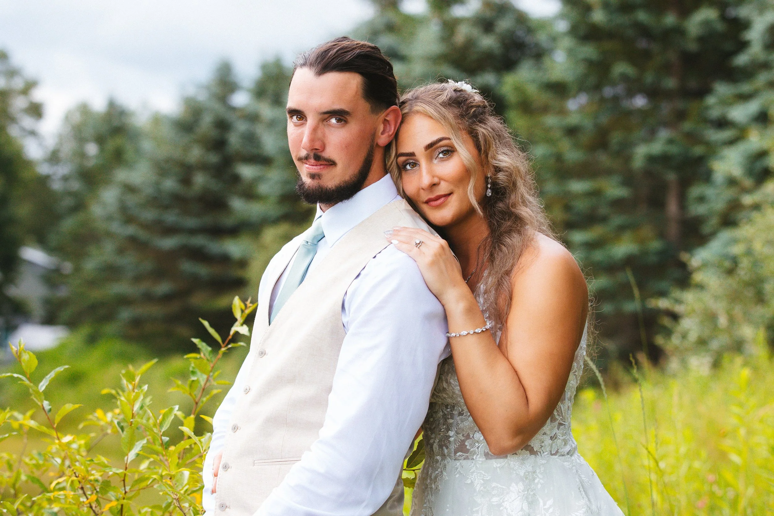 A newlywed couple outdoors with greenery and trees in the background, the groom wearing a cream vest and light blue shirt, and the bride in a white lace wedding dress with wavy hair, smiling and embracing.