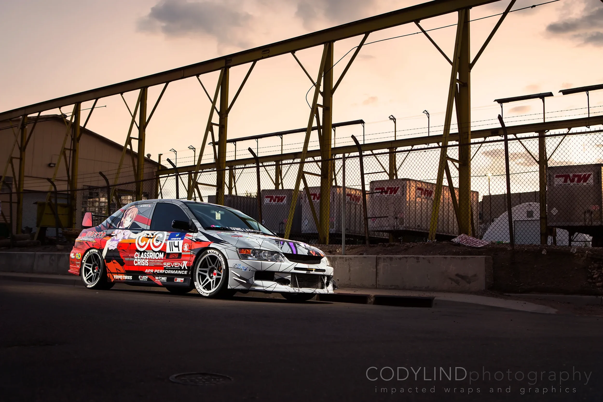 A race car with anime-style graphics and various sponsor stickers parked on a city street at sunset, behind a chain-link fence with trailers labeled 'TMW' in the background.