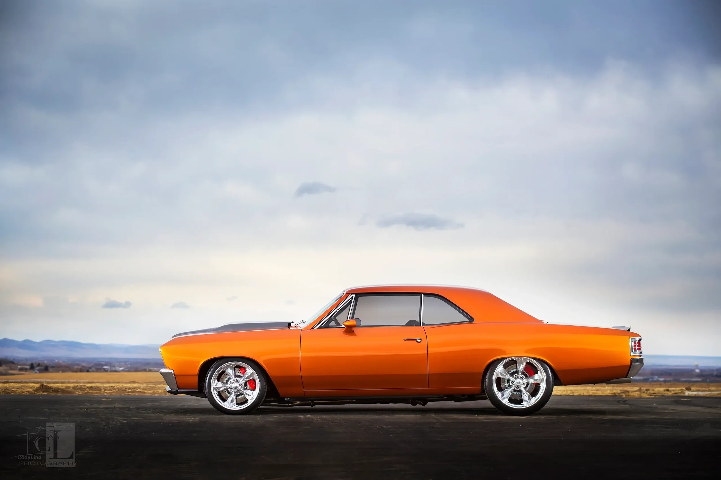 Orange vintage car parked on a paved road with a cloudy sky and distant mountains in the background.
