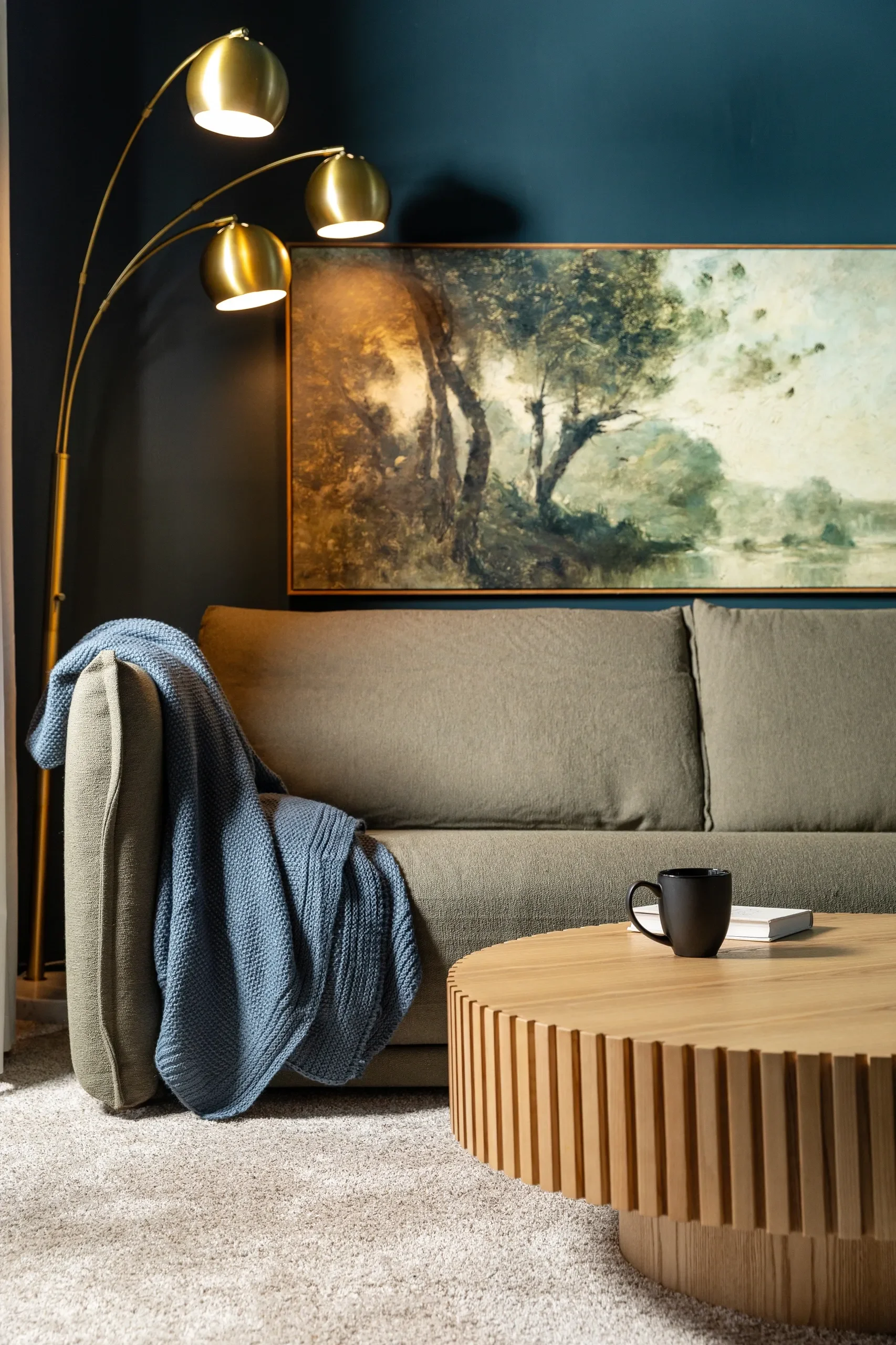 Living room with a gray sofa, a blue blanket draped over the armrest, a wooden coffee table with a black mug and a white book, a large landscape painting, and a floor lamp with three gold-colored shades.