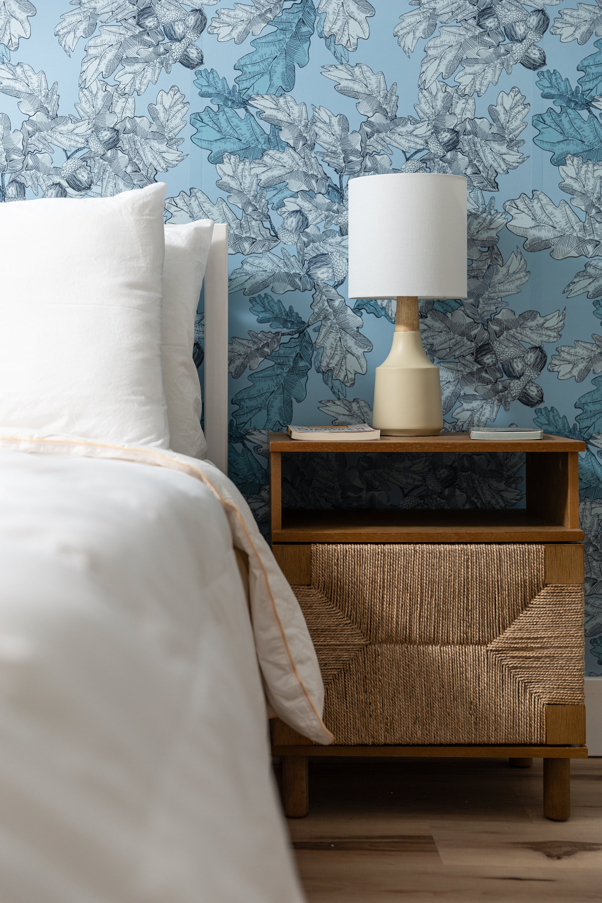 A bedroom corner with a white bed and pillows, a wooden nightstand with a woven front drawer, a ceramic lamp with a white shade, and a blue leafy patterned wallpaper background.
