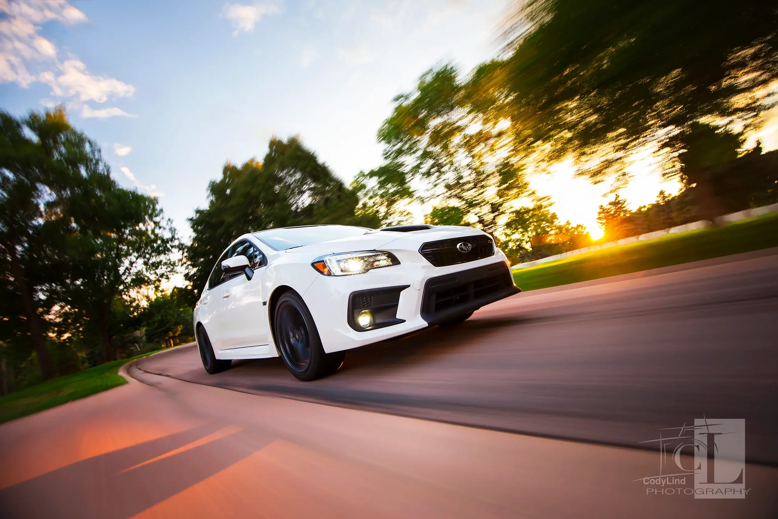 A white Subaru car driving on a curved road during sunset with trees in the background.