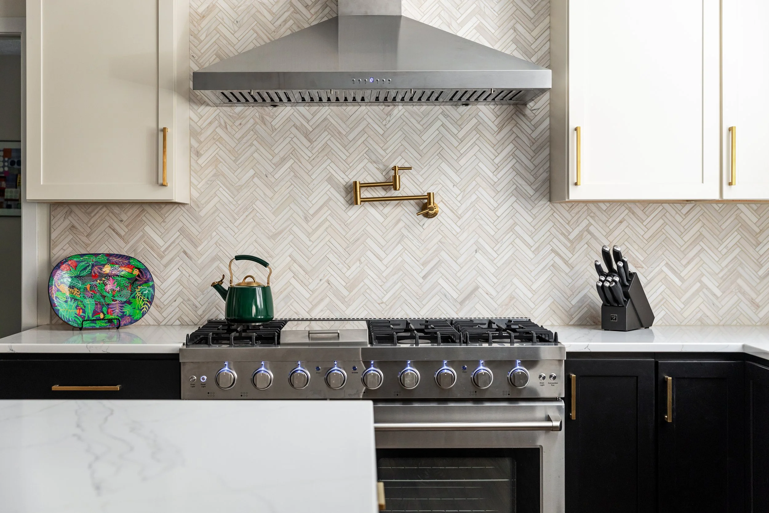 Modern kitchen with a stainless steel stove, white marble countertops, white cabinets with gold handles, a colorful decorative plate, a green kettle, and a knife block. The backsplash has a herringbone pattern.