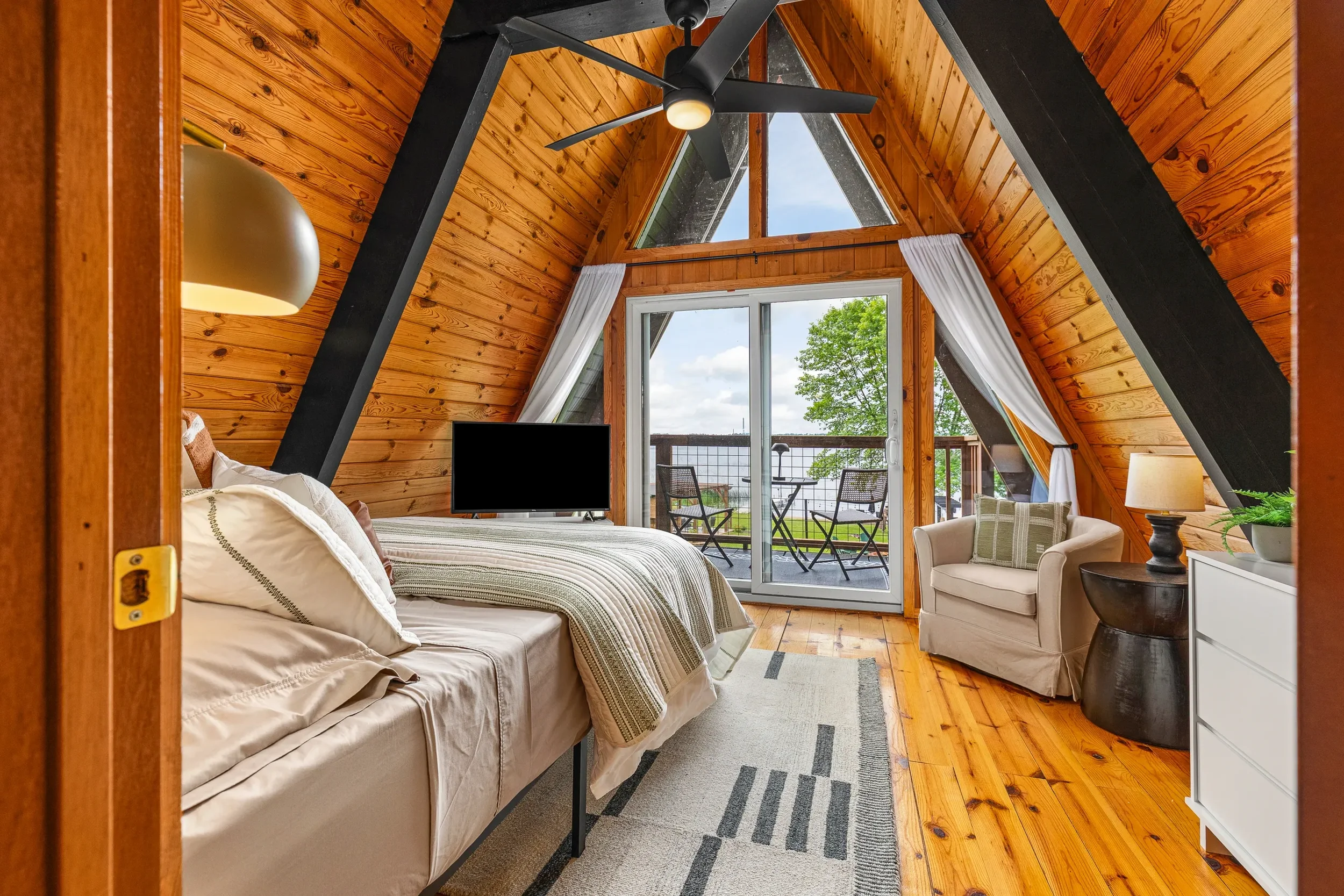 A cozy bedroom with wooden walls and ceiling, a large bed with white linens, a small balcony with outdoor seating, and a view of trees and water outside.