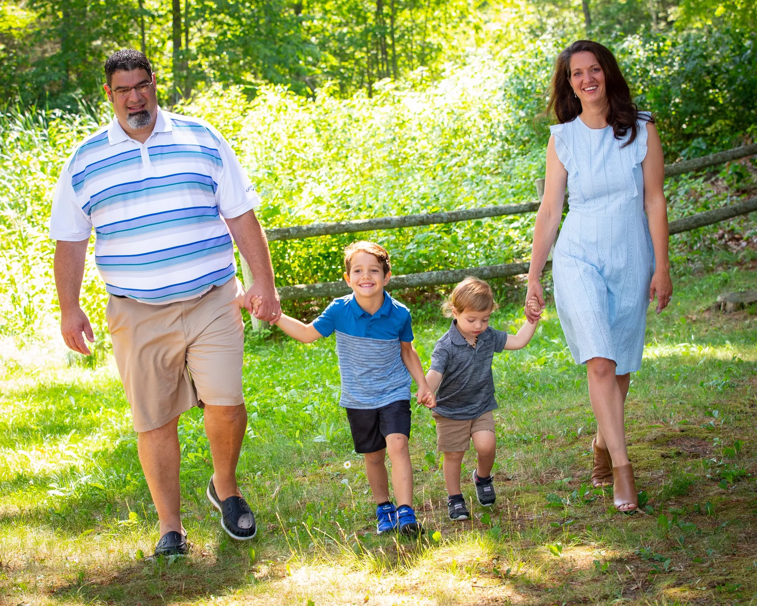 A happy family of four, with two adults and two children, walking hand in hand through a lush green park on a sunny day.