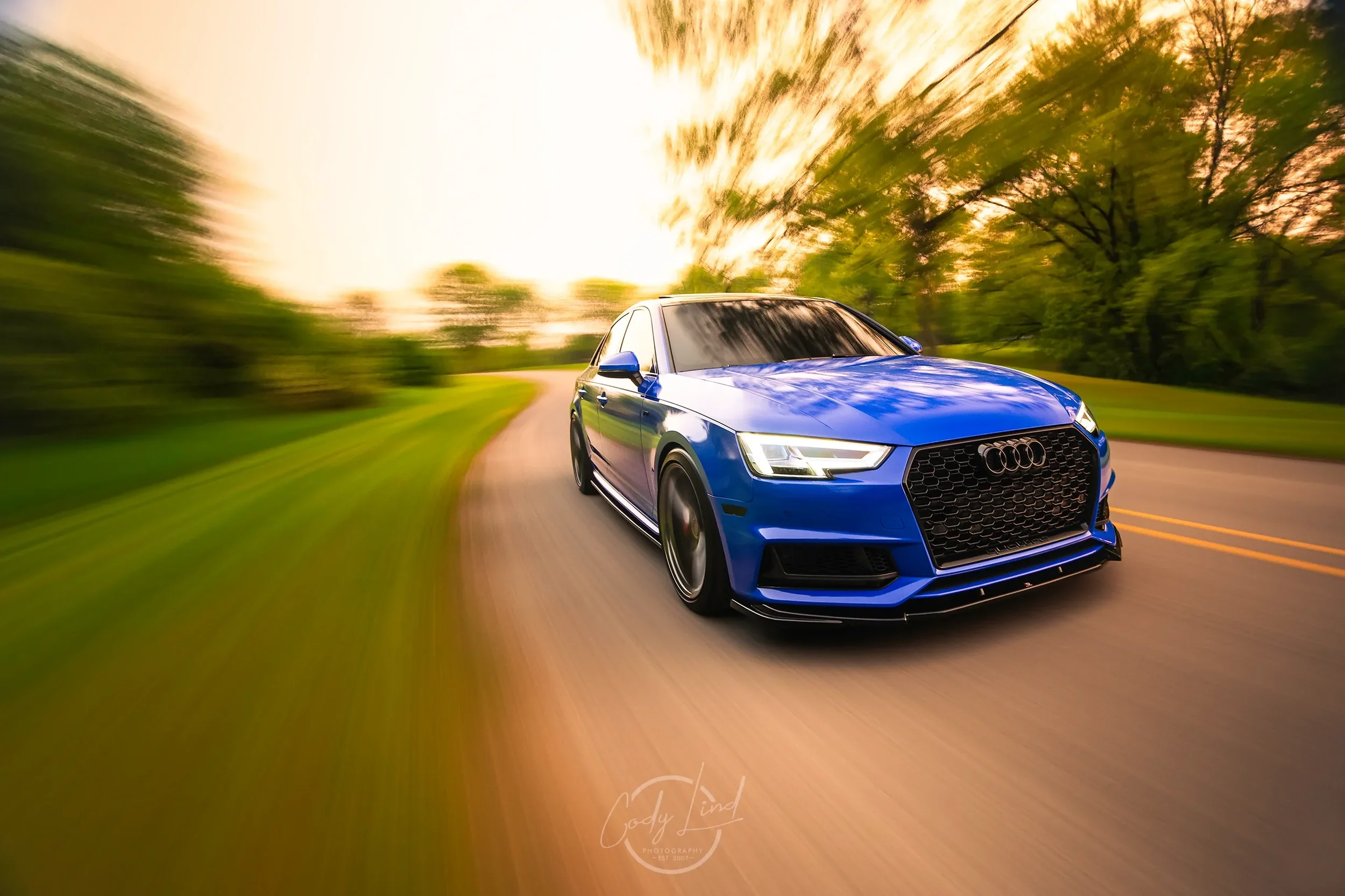 Blue Audi sedan driving on a winding road surrounded by green trees during sunset with motion blur effect.