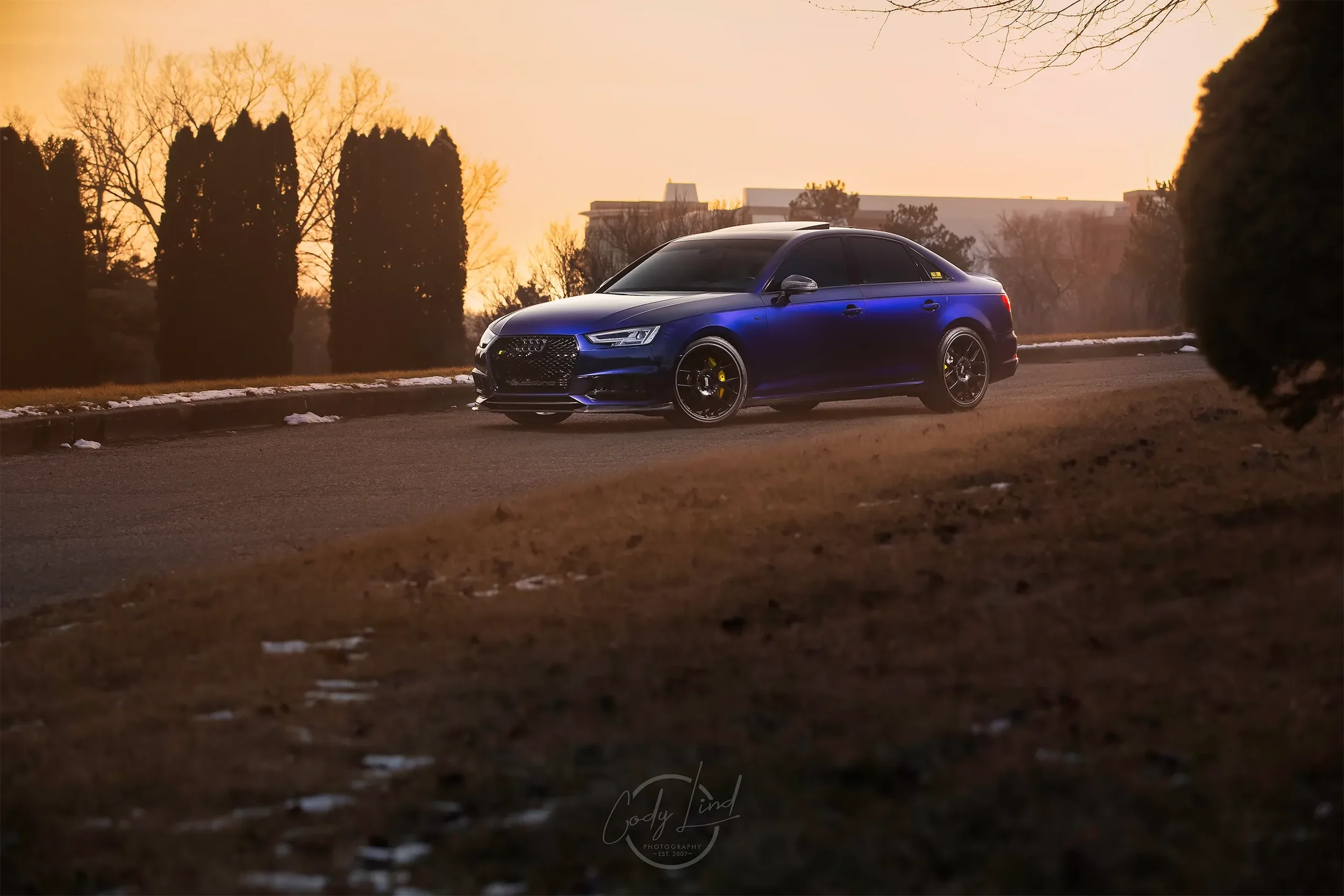 A blue sports car parked on a quiet road during sunset, with trees and buildings in the background.