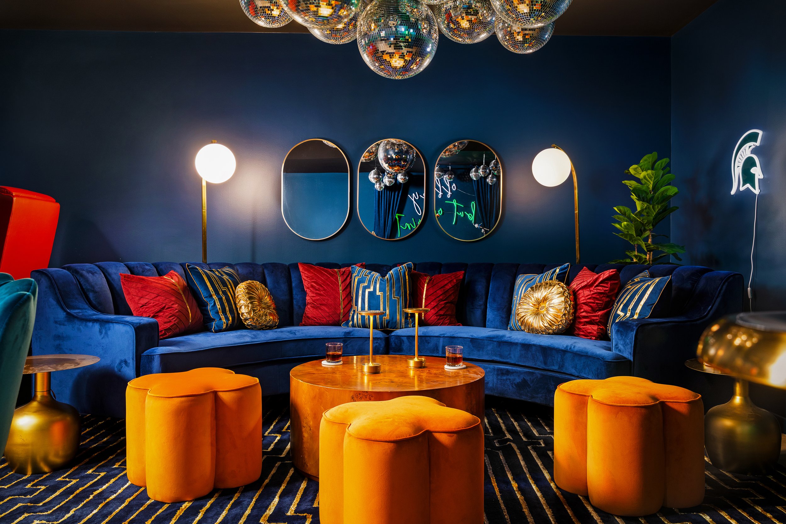 A vibrant living room with a deep blue curved velvet sofa adorned with gold, red, and striped pillows, a wooden coffee table with gold candles, and orange ottomans. The walls are dark blue with three mirrors and modern lighting fixtures, including gl