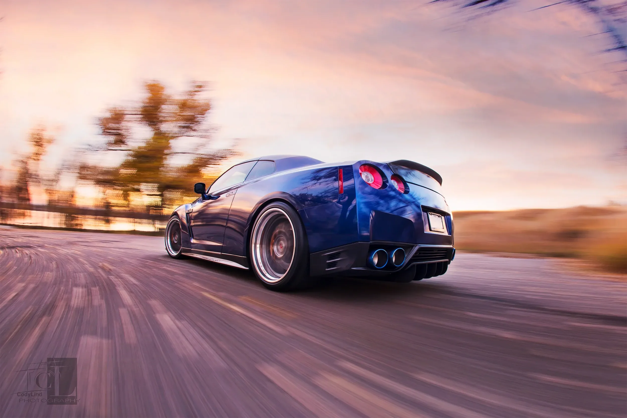 A blue sports car driving on a road during sunset, with motion blur indicating high speed and a scenic background of trees and a colorful sky.