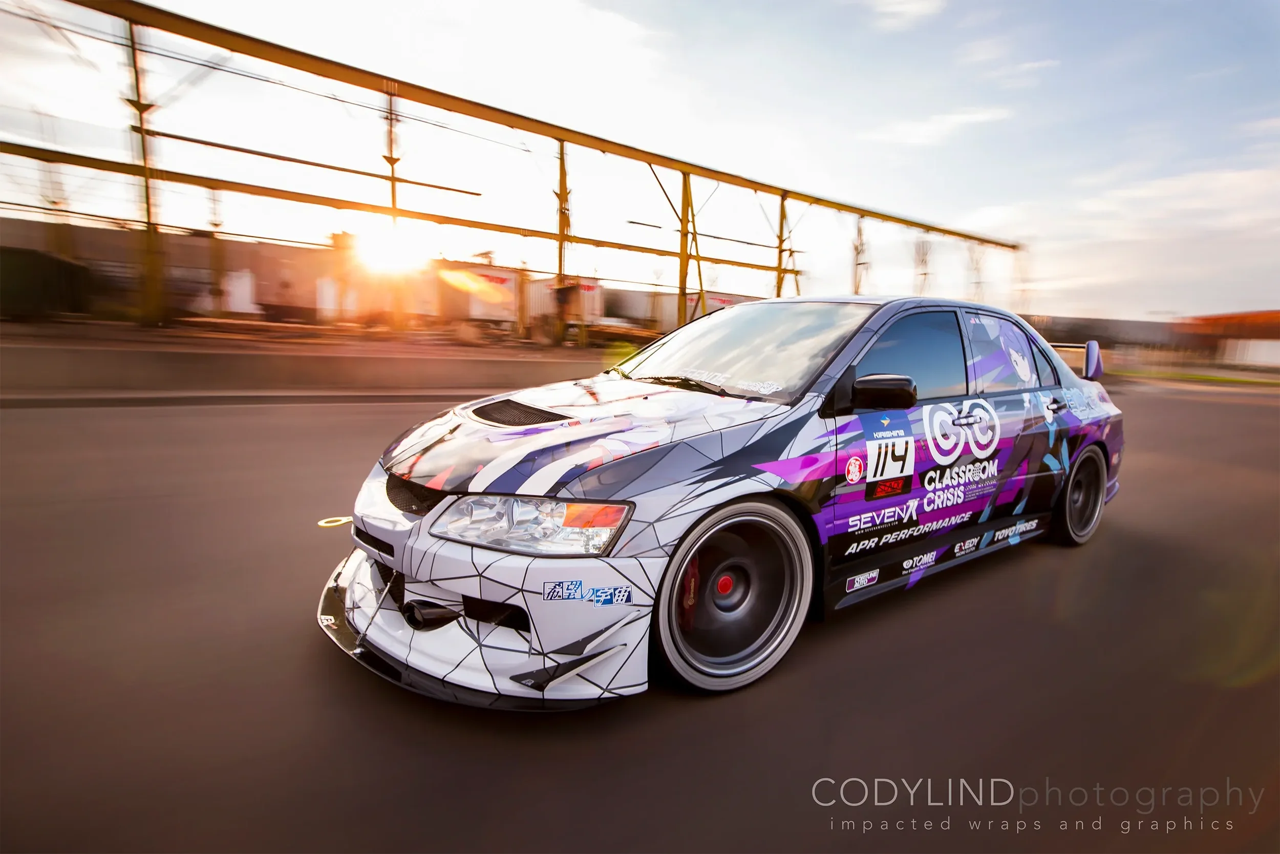 A racecar with custom graphics driving on a road during sunset, featuring a front splitter, aerodynamic body kit, and decals including the number 114 and event name 'Seven7 Classroom Crisis'.