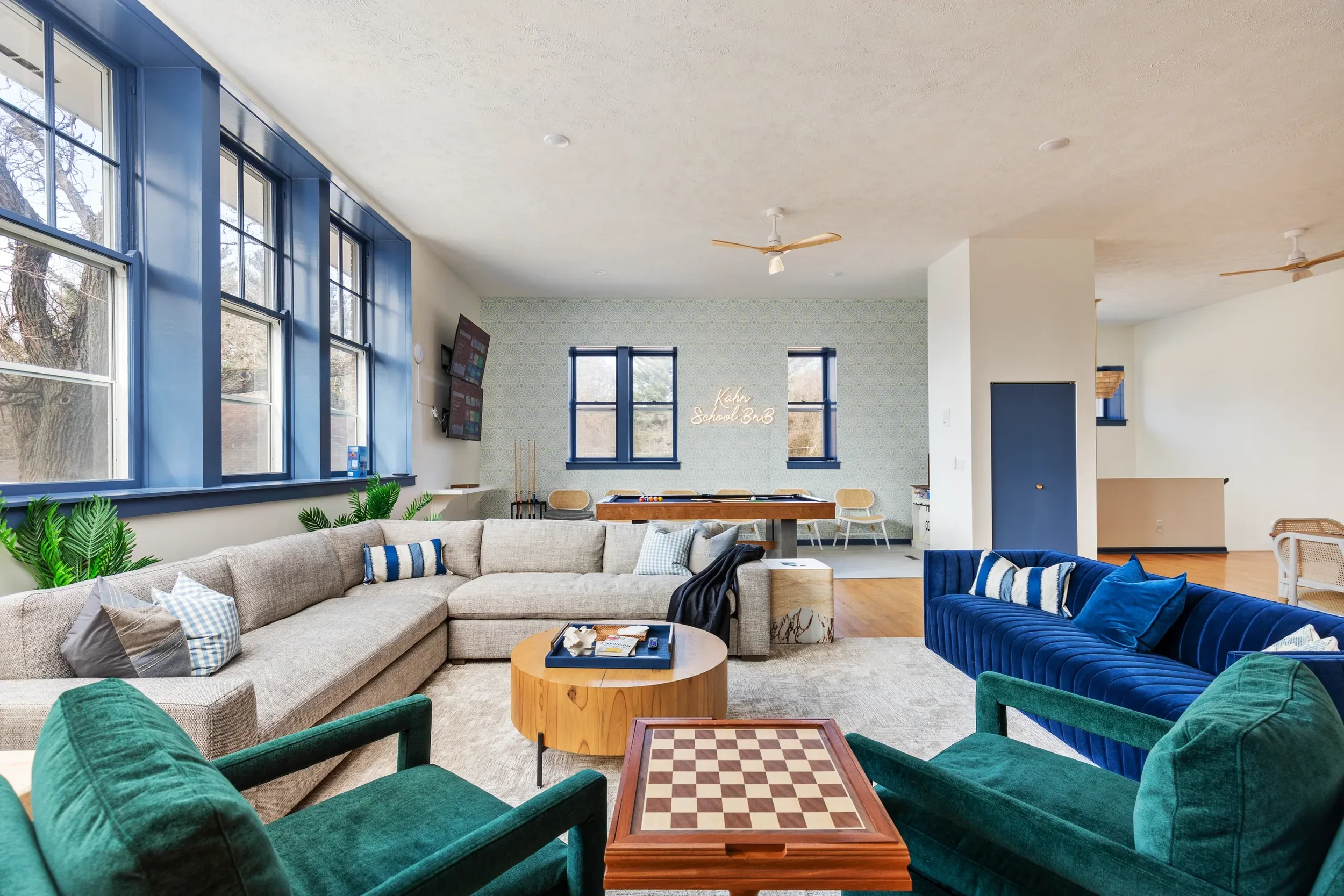 Living room with large window, beige sectional sofa, green armchair, blue velvet sofa, wooden coffee table, chessboard, plants, wall with neon sign saying 'Koko School BnB', TV, and backgammon table.