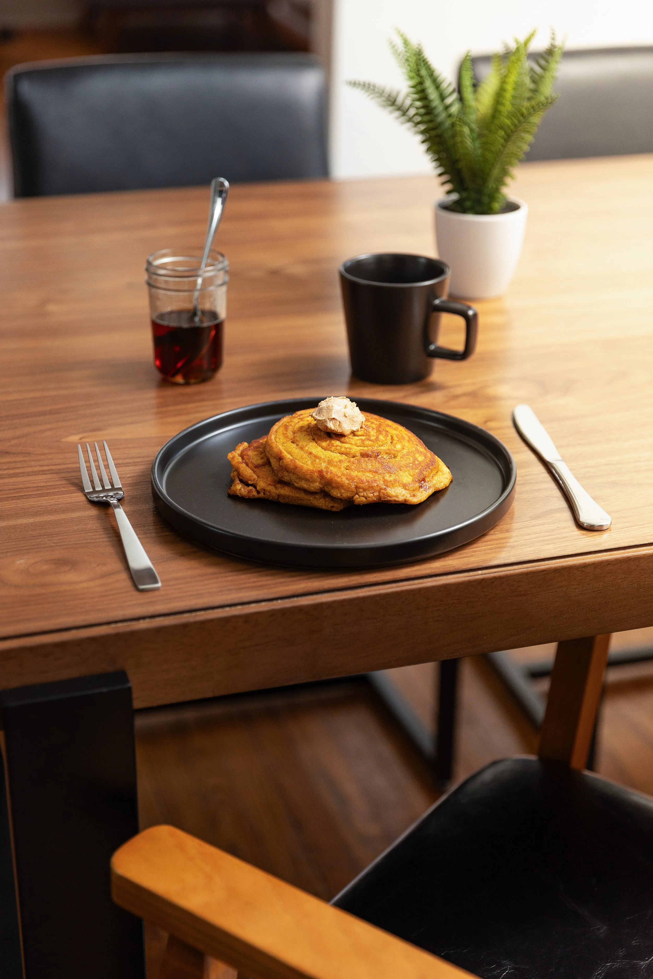 A table with a plate holding a cinnamon roll with icing on top, a jar of jam with a spoon inside, a black coffee mug, a white potted fern plant, a black plate, a fork on the left, and a knife on the right, with two black chairs and wooden flooring in